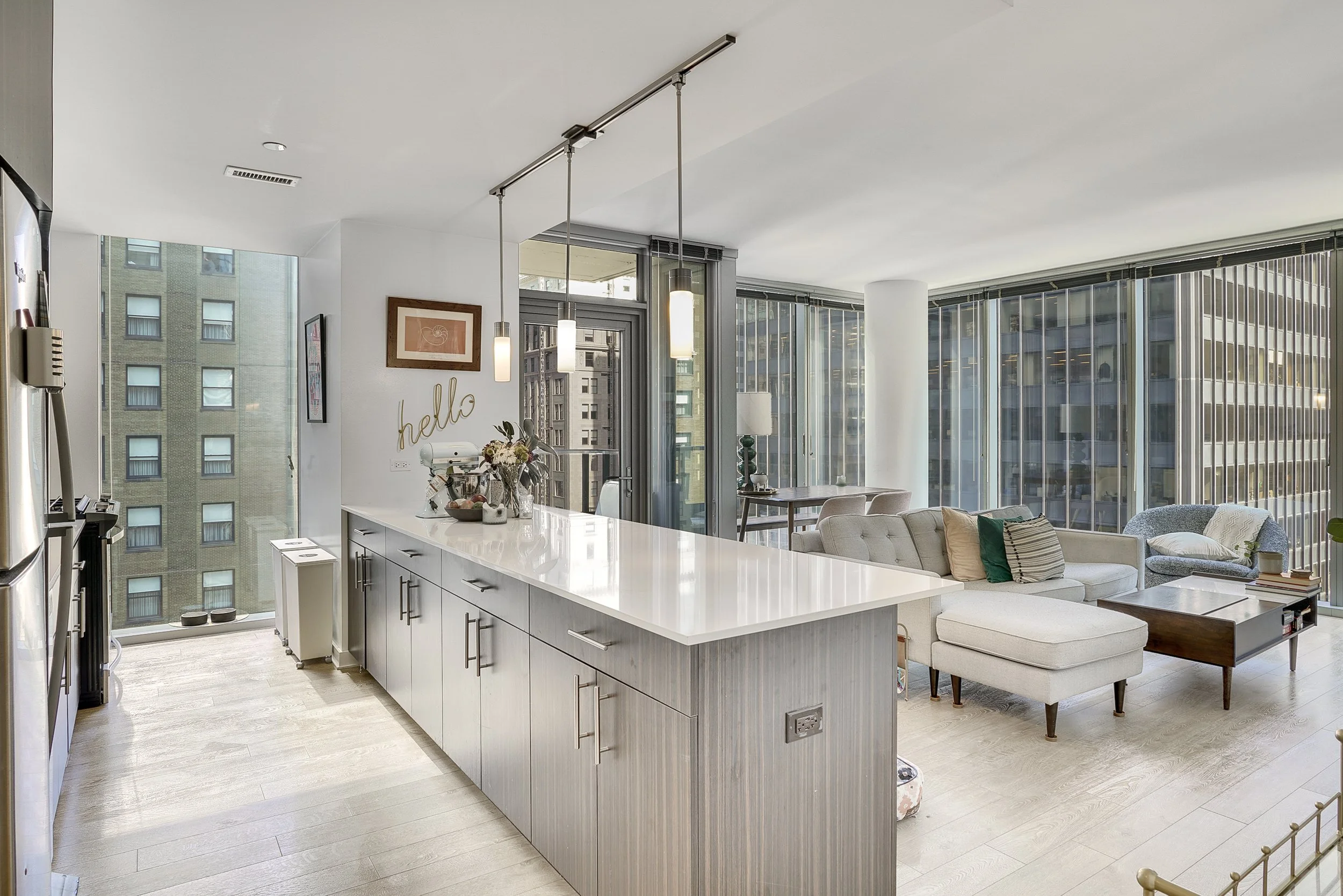 Modern open-concept living room and kitchen with large windows, light-colored furniture, and urban city views.