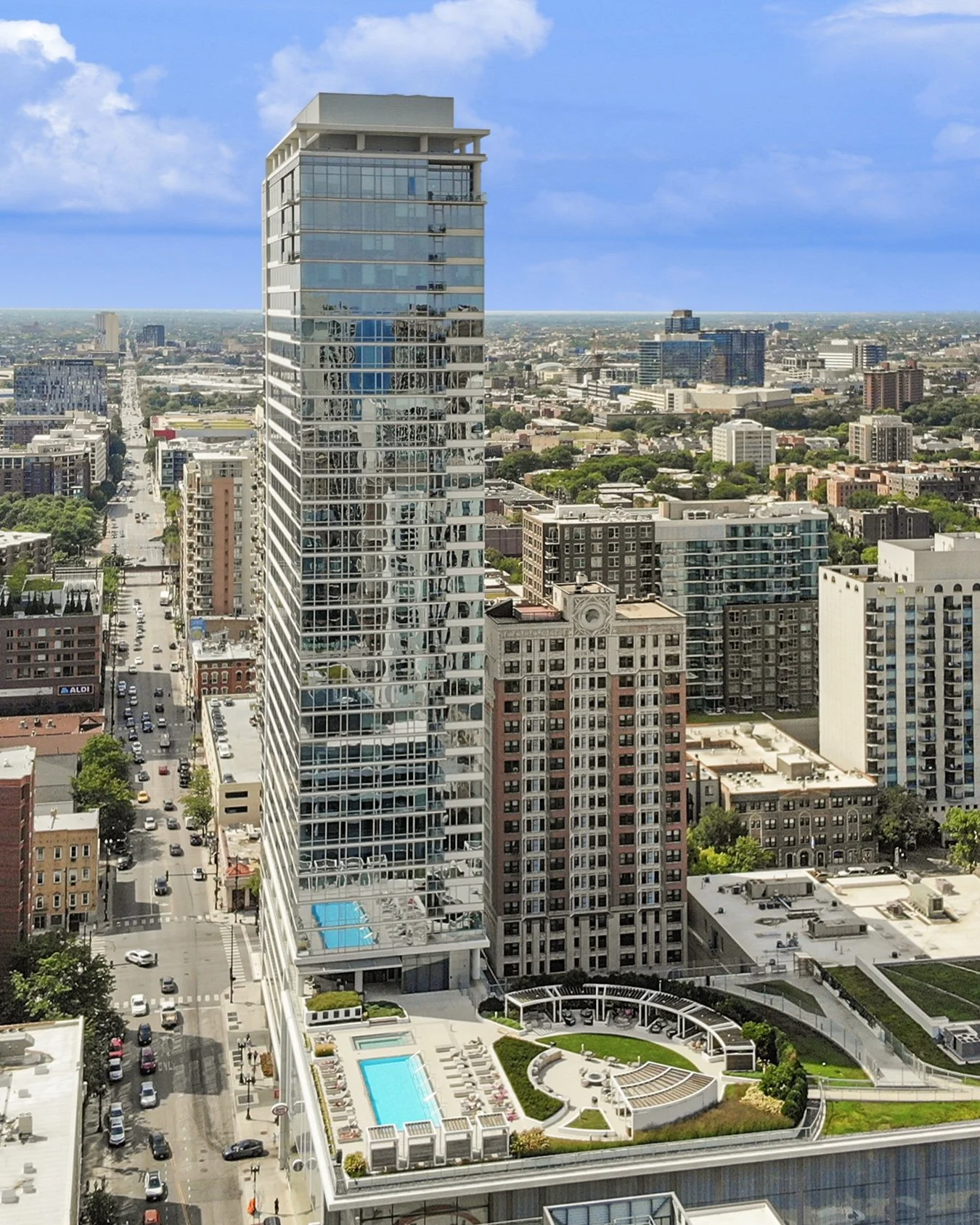 A tall modern glass skyscraper with balconies and a rooftop pool, situated among other buildings in a cityscape with a clear blue sky.
