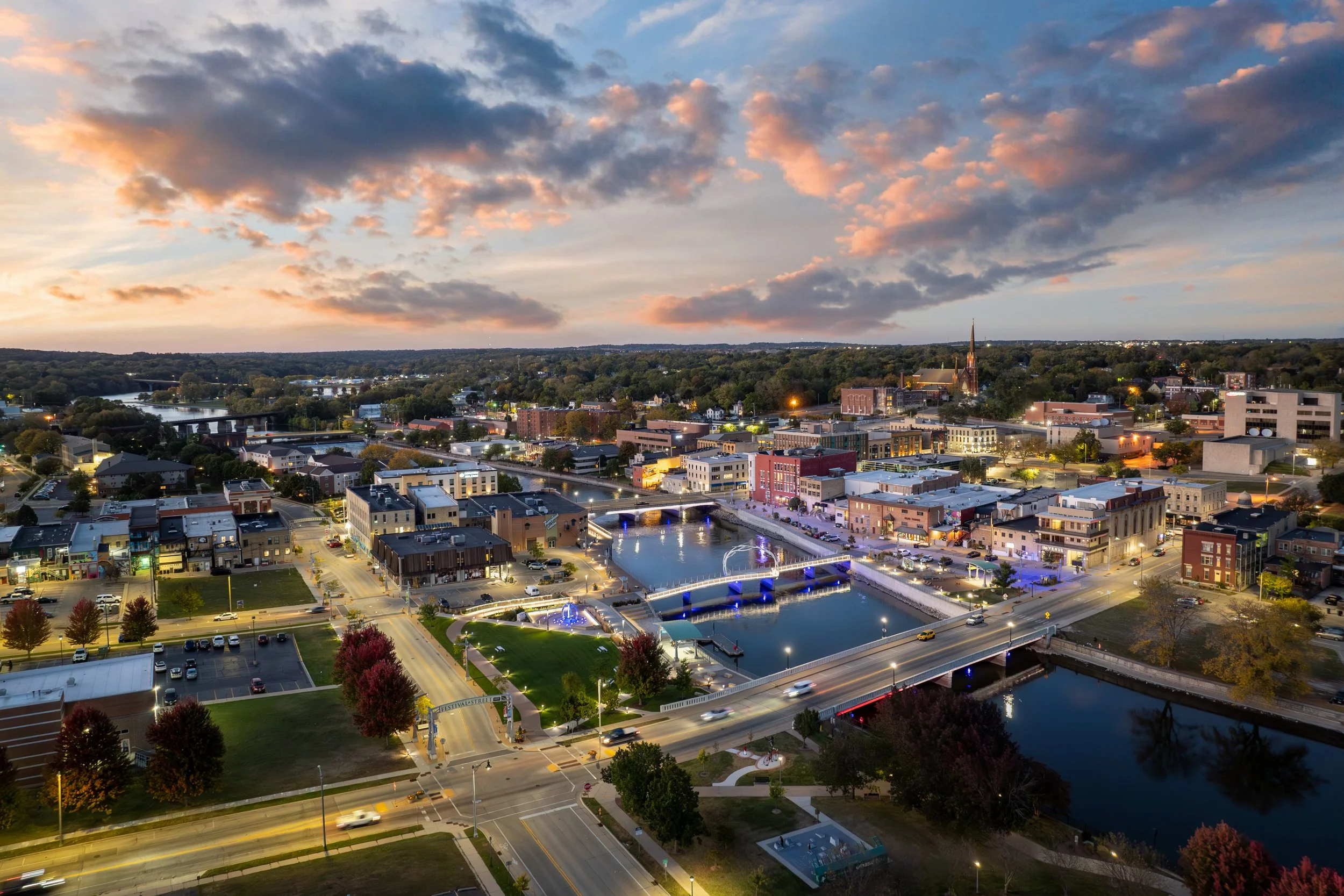 Aerial view of a city at dusk with buildings, a river, and bridges, with a colorful sky and scattered clouds.
