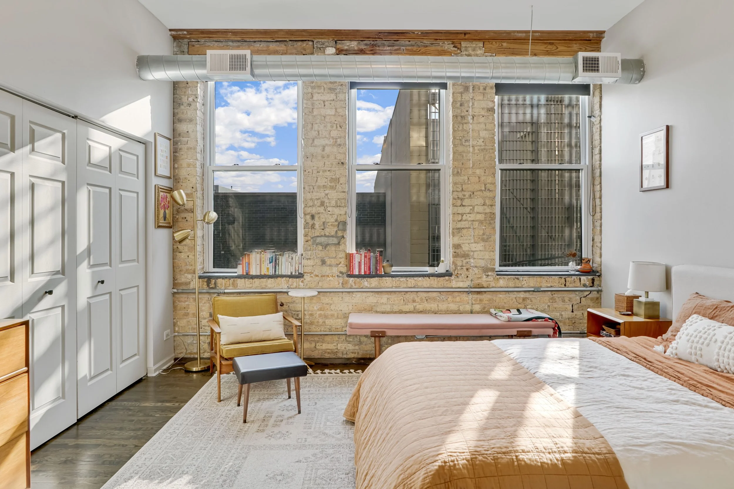 Bright bedroom with exposed brick walls, large windows, a bed with beige bedding, a yellow armchair, a small ottoman, and a modern standing lamp.