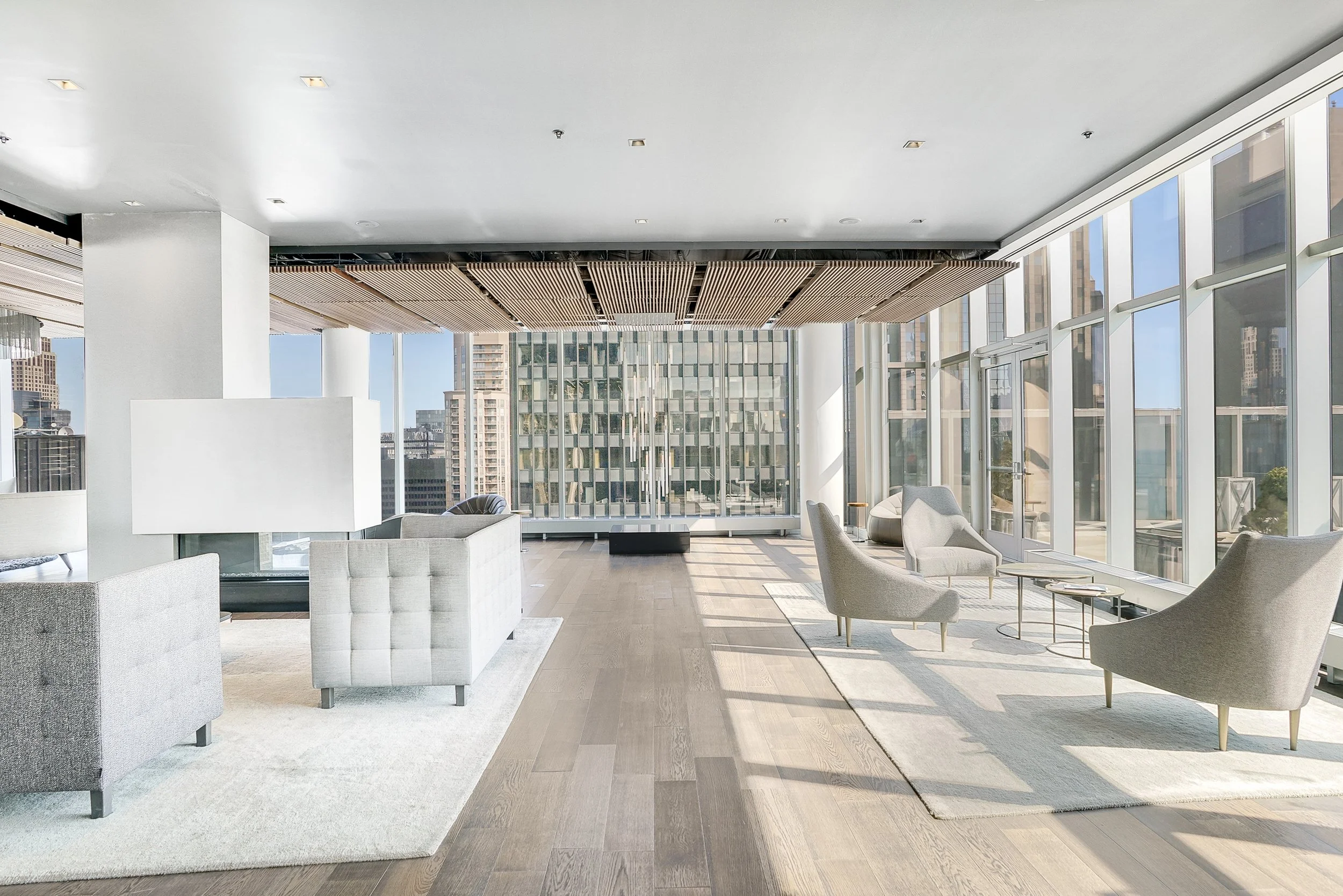 Modern lounge with beige armchairs and sofas, large windows overlooking a city skyline, light wooden floors, and a white area rug.