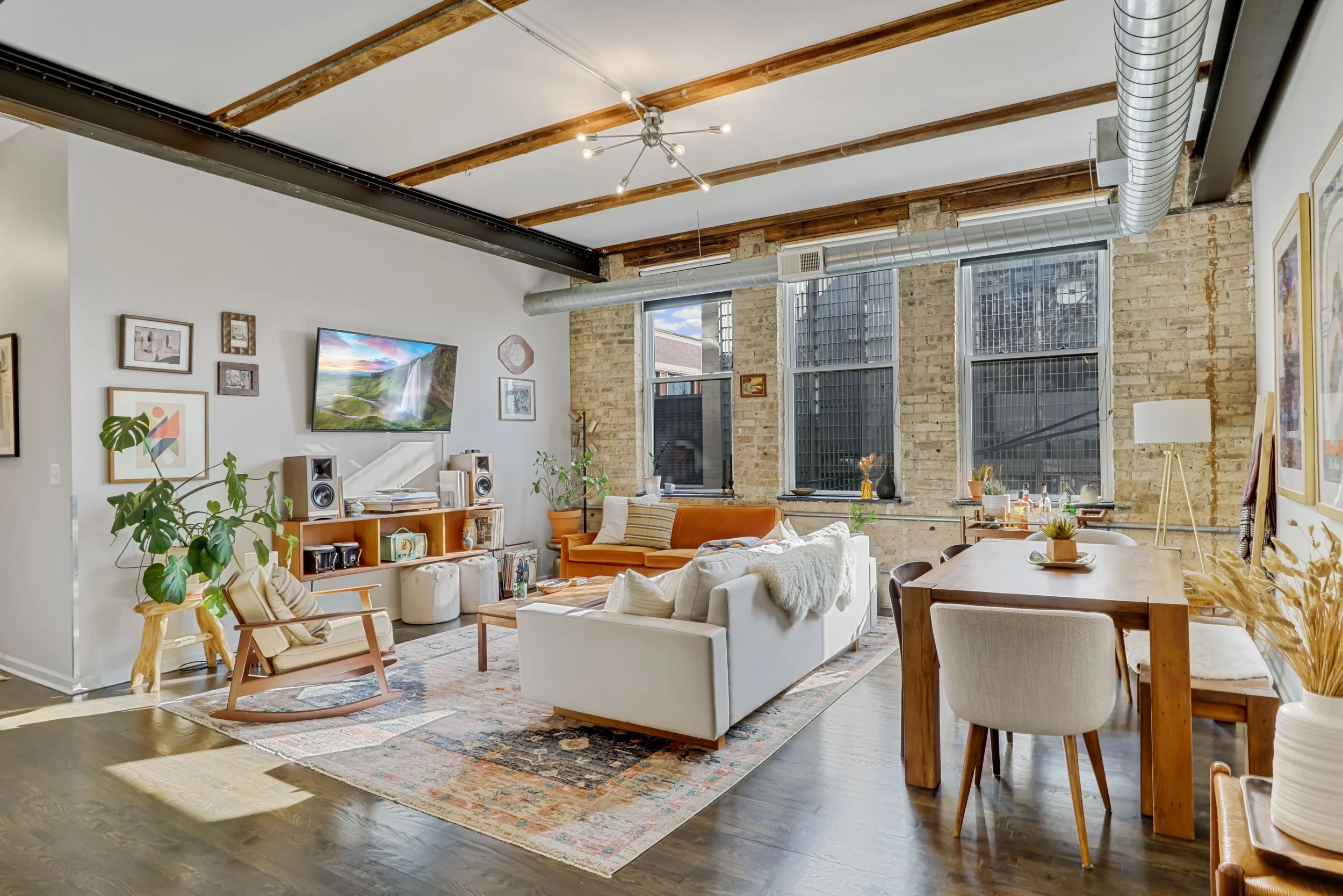 Spacious living room with white and orange sofas, wooden table, colorful wall art, large windows, exposed brick wall, and ceiling with wooden beams and modern lighting fixture.