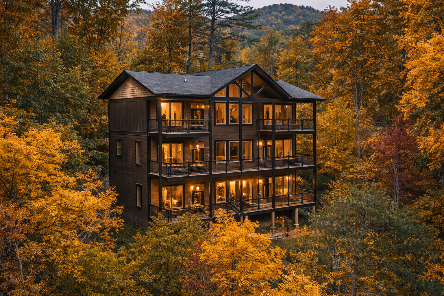 A modern, multi-story wooden house with large windows and balconies is nestled among colorful fall trees in a forest, illuminated from within at dusk.