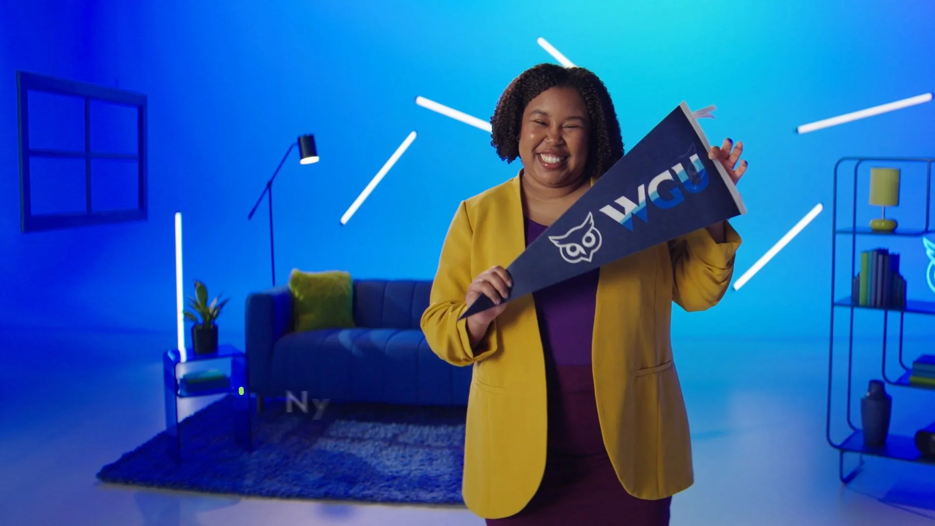 Smiling woman holding WGU sign in modern living room with blue lighting, sofa, bookshelf, and decorative lighting.