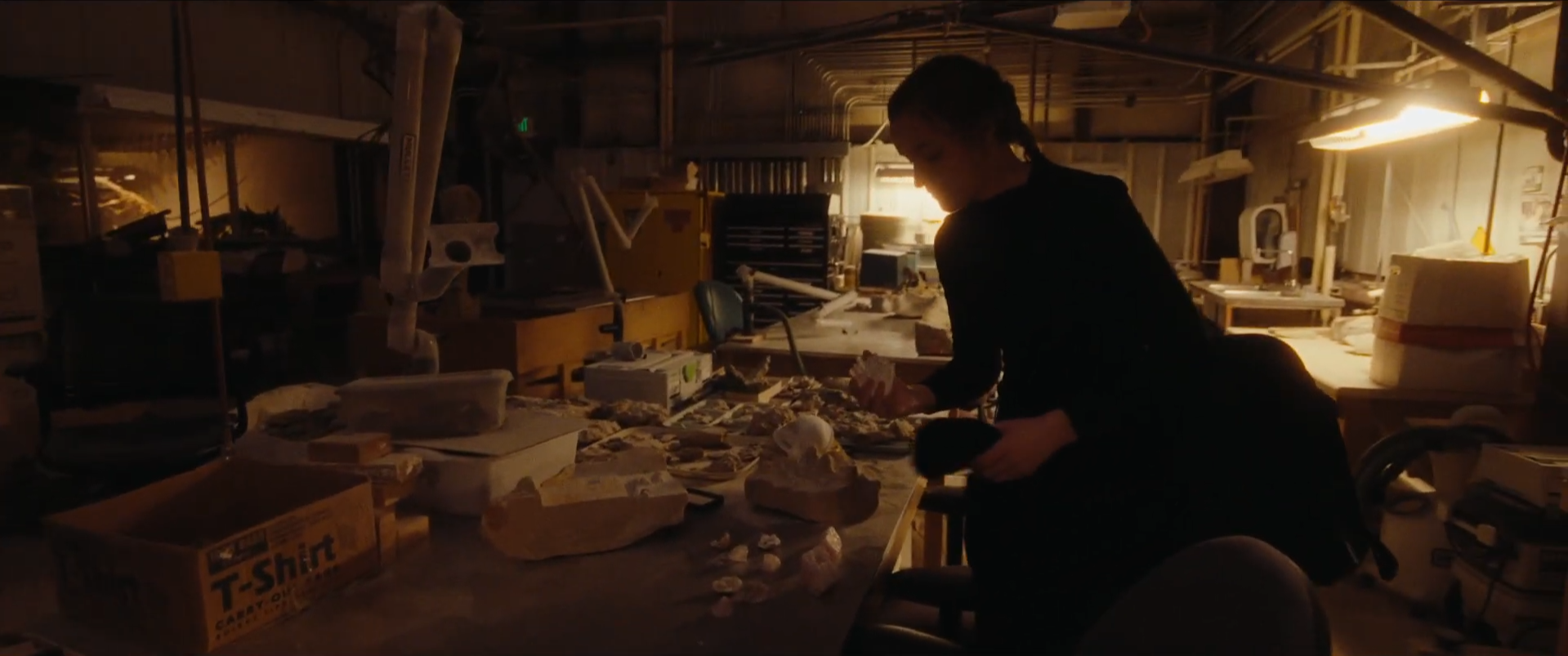 A person working in a dimly lit workshop or studio, surrounded by various materials and tools, with warm lighting illuminating their workspace.