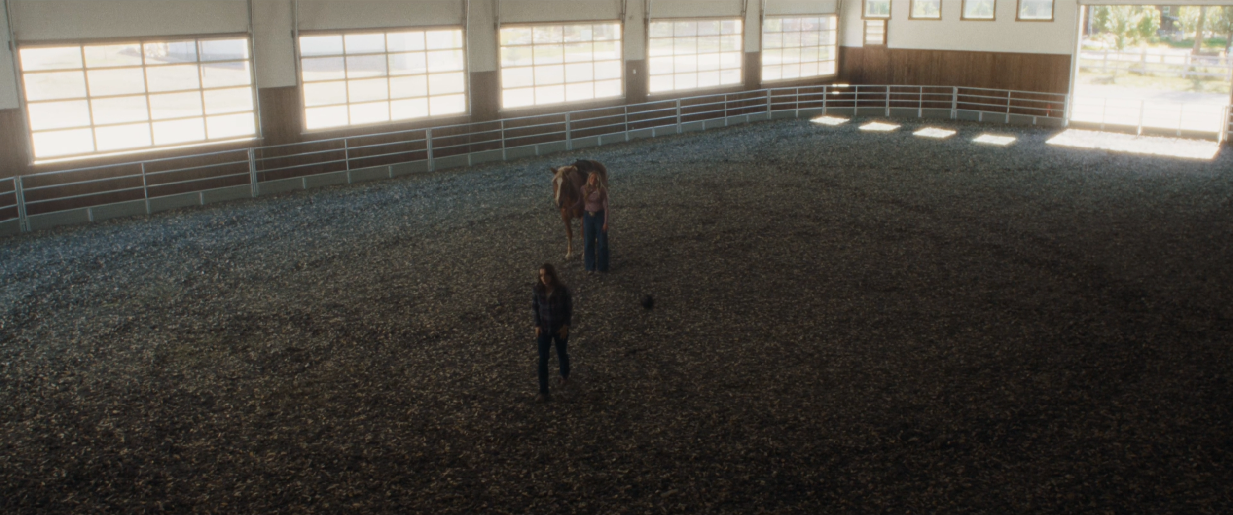 An indoor riding arena with large windows, a woman walking on the sandy floor, and a person standing near a brown horse.