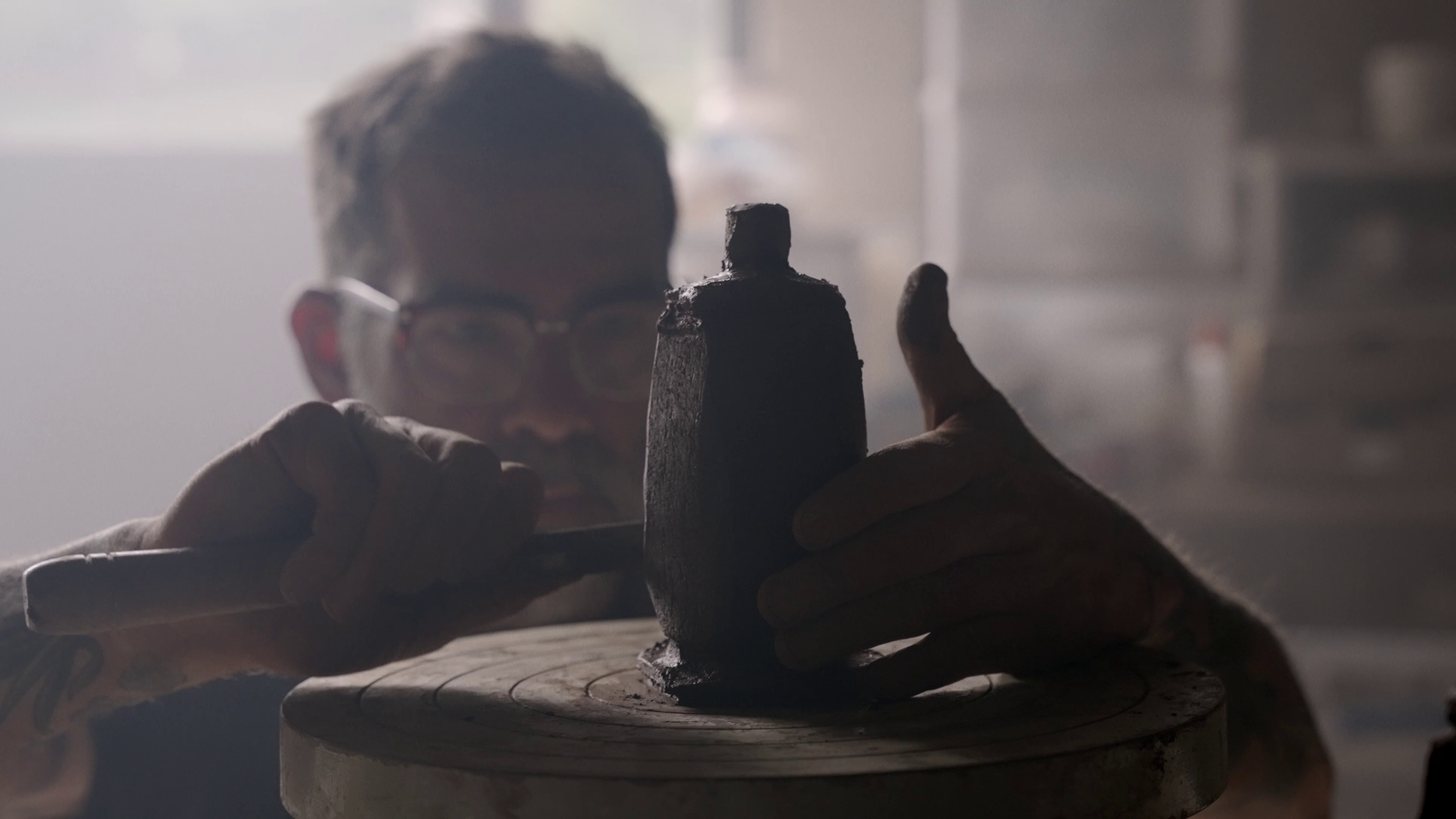 Person holding a small dark sculpture or object, working on a pottery wheel in a studio or workshop with shelves in the background.