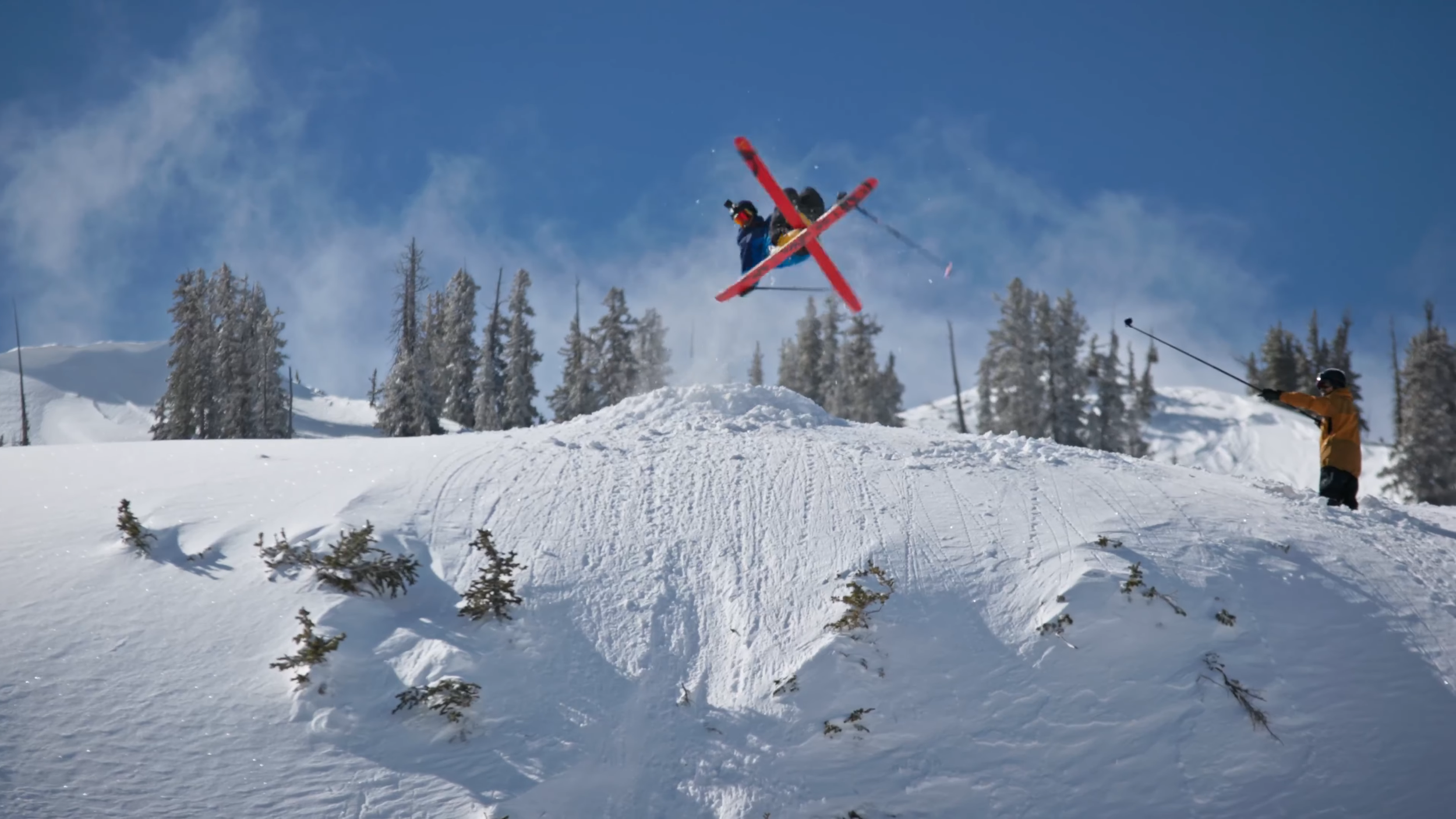 A skier airborne during a jump on a snowy mountain, with a person in an orange jacket holding a ski pole, and snow-covered trees in the background.