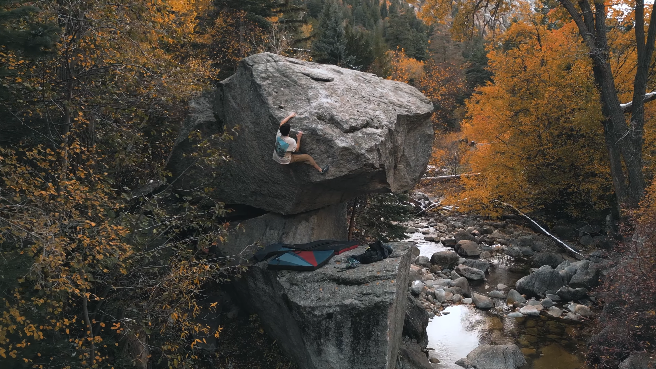 A person bouldering on a large rock formation next to a creek in a forest with autumn-colored trees.