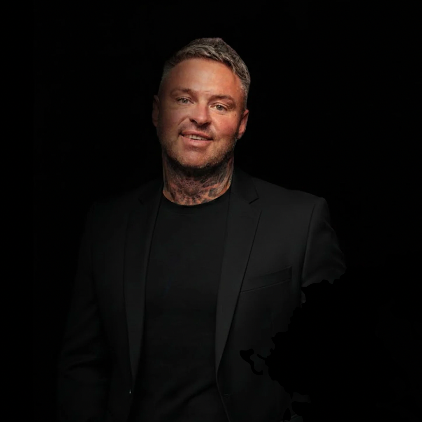 A man with short, grayish hair and tattoos on his neck, wearing a black blazer and a black shirt, smiling against a black background.