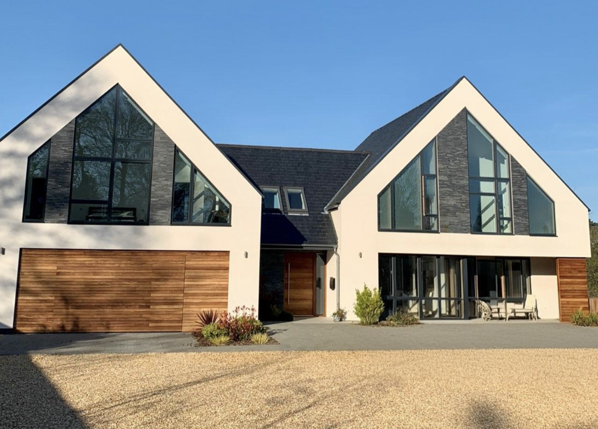 Modern house with large glass windows, white walls, dark roof, and wooden accents, situated in a sunny outdoor setting.