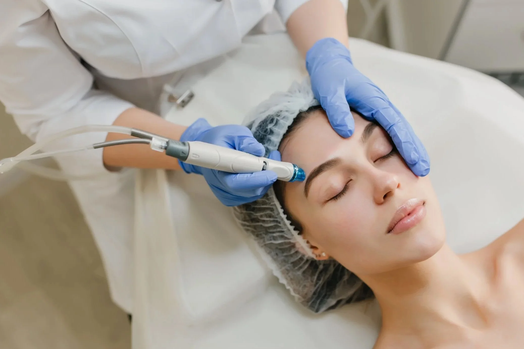 A woman receiving a facial treatment with a laser device, lying on a treatment bed with eyes closed, wearing a hair cover, with a technician in blue gloves performing the procedure.