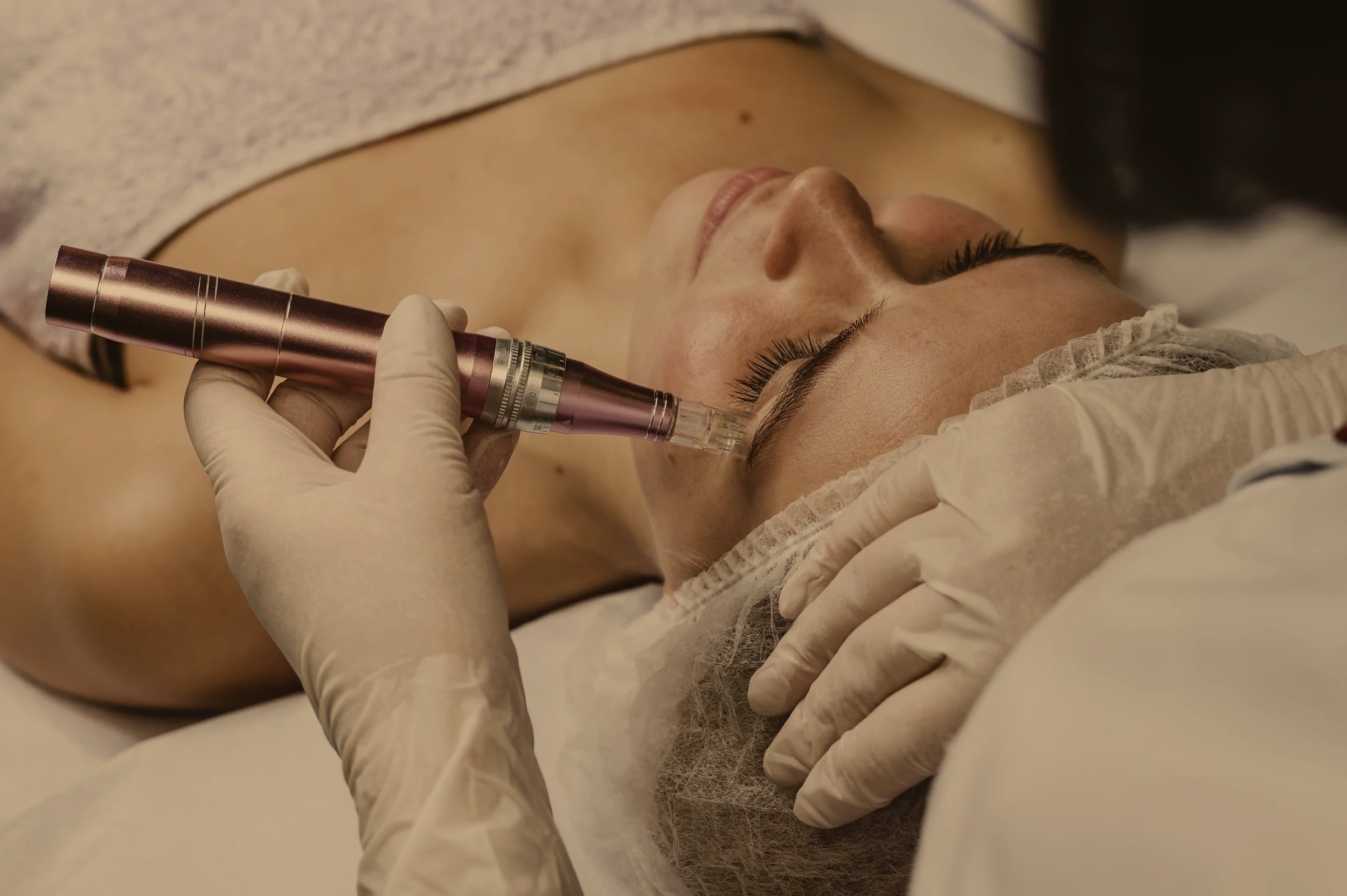 A woman lying down with her eyes closed, receiving a cosmetic or dermatological treatment with a handheld device from a professional wearing gloves, in a clinical setting.