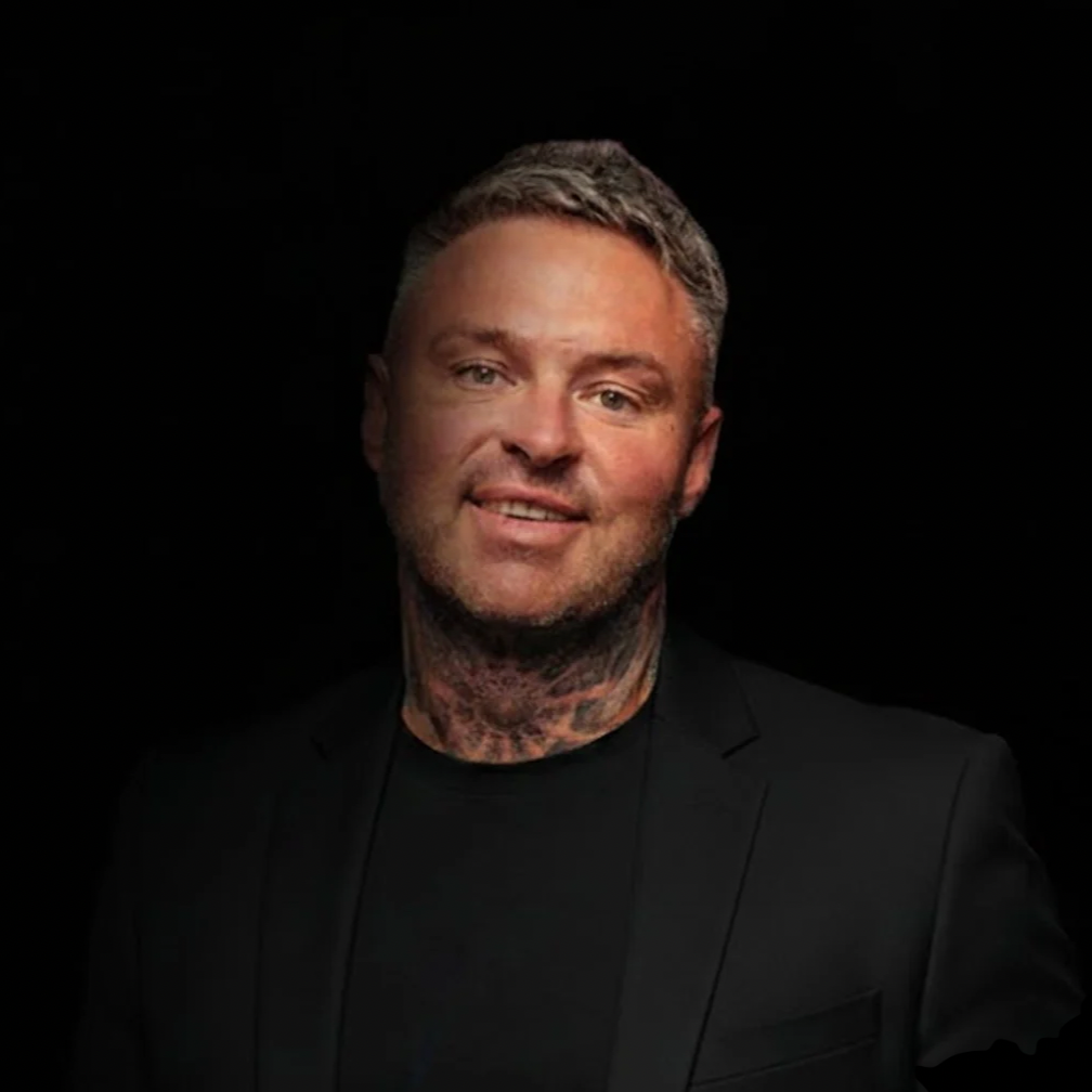 A portrait of a man with short blonde hair, tattoos on his neck, wearing a black blazer and black shirt against a dark background.