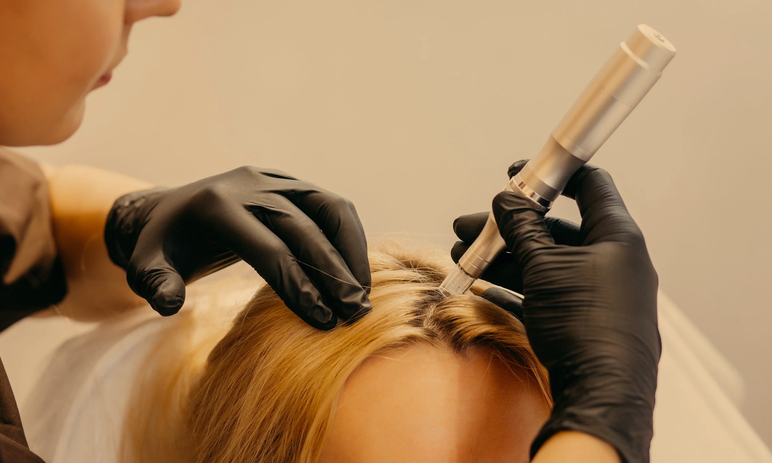 A person in black gloves performs a cosmetic tattoo procedure on a woman's eyebrows using a handheld tattooing device.