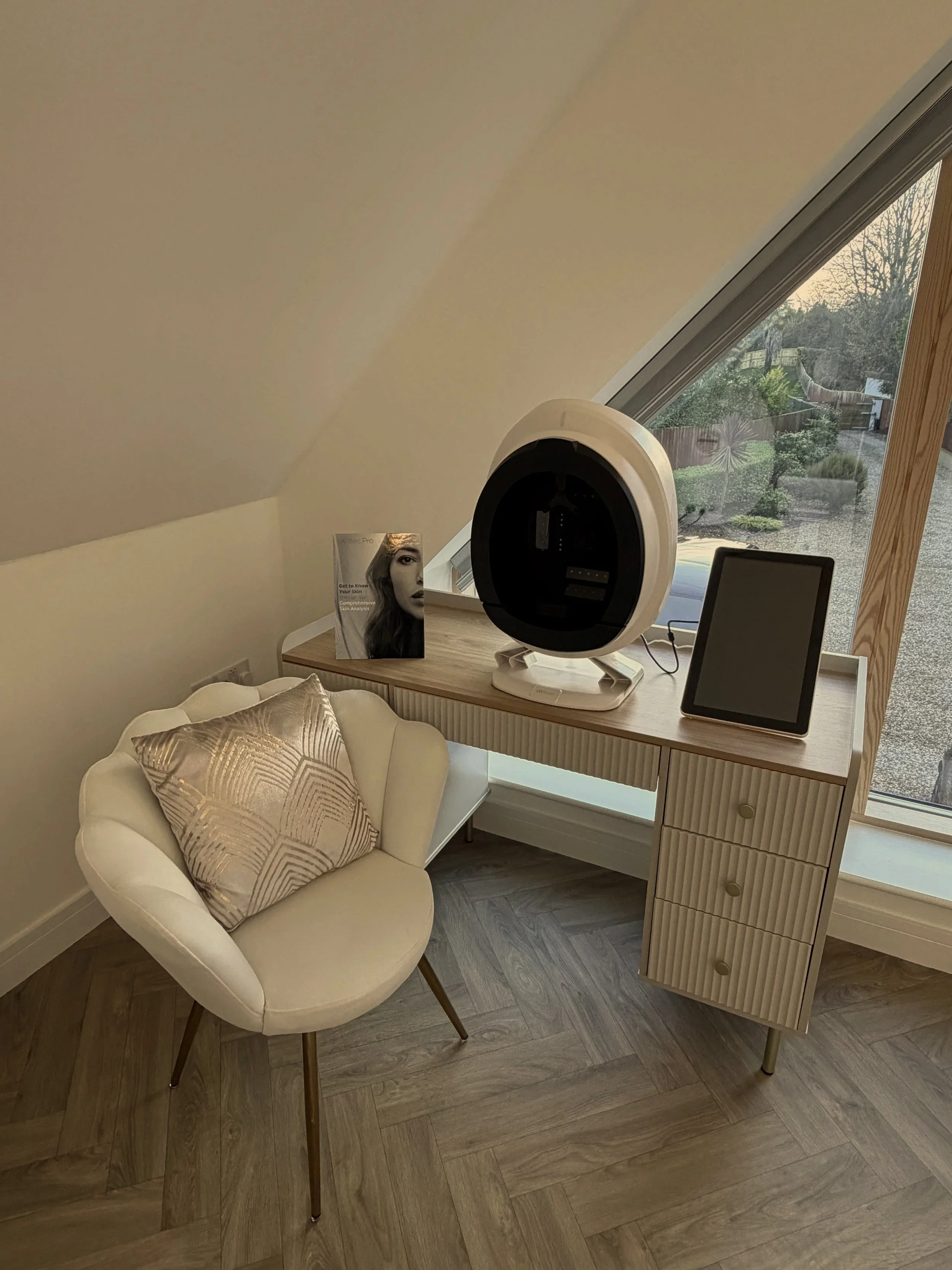 A cozy home office corner with a white chair, a wooden desk, a futuristic device, a tablet, and a magazine, near a large window with a slanted ceiling.
