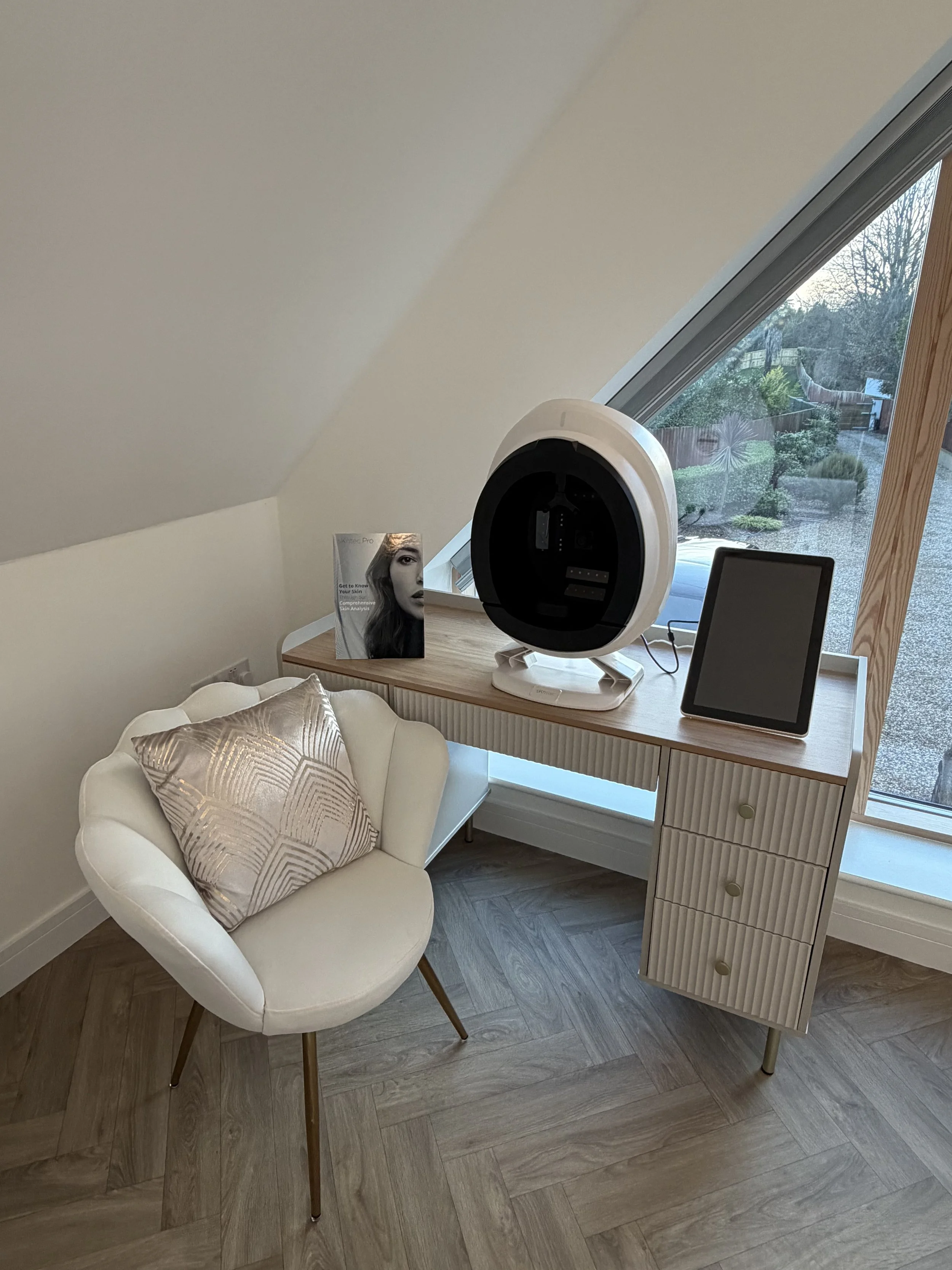 A modern home office corner with a white armchair and a cute throw pillow, a wooden desk with an abstract design, a tablet, a sleek black and white device, and a magazine. The setup is near a large triangular window overlooking a garden.