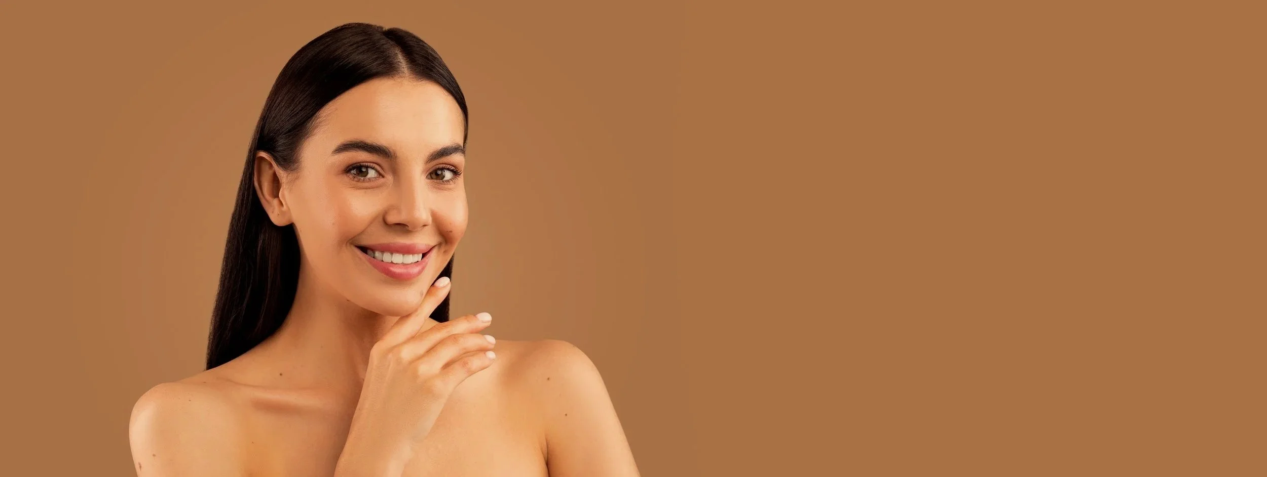 Smiling young woman with long dark hair, touching her chin, with a warm brown background.