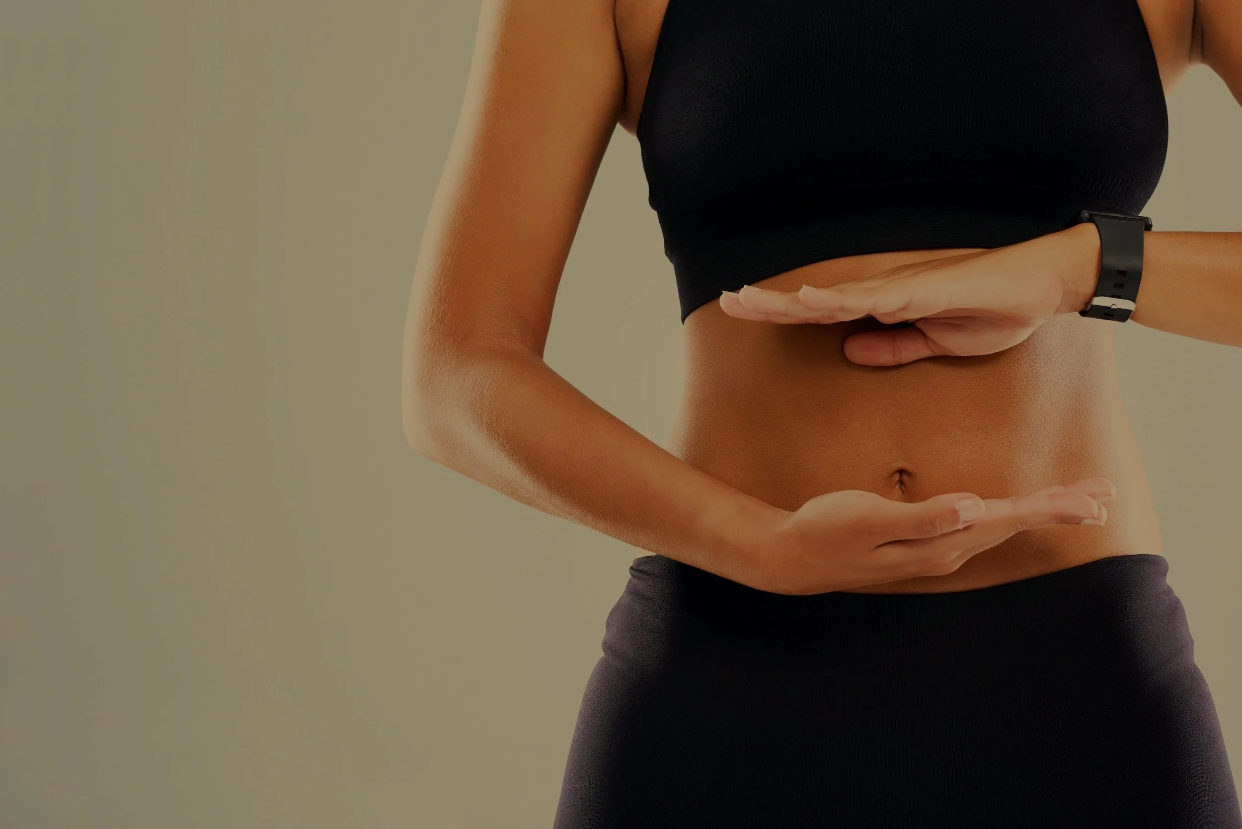 Close-up of a woman's midsection with hands making a frame around her stomach, wearing a black sports bra and black workout pants.
