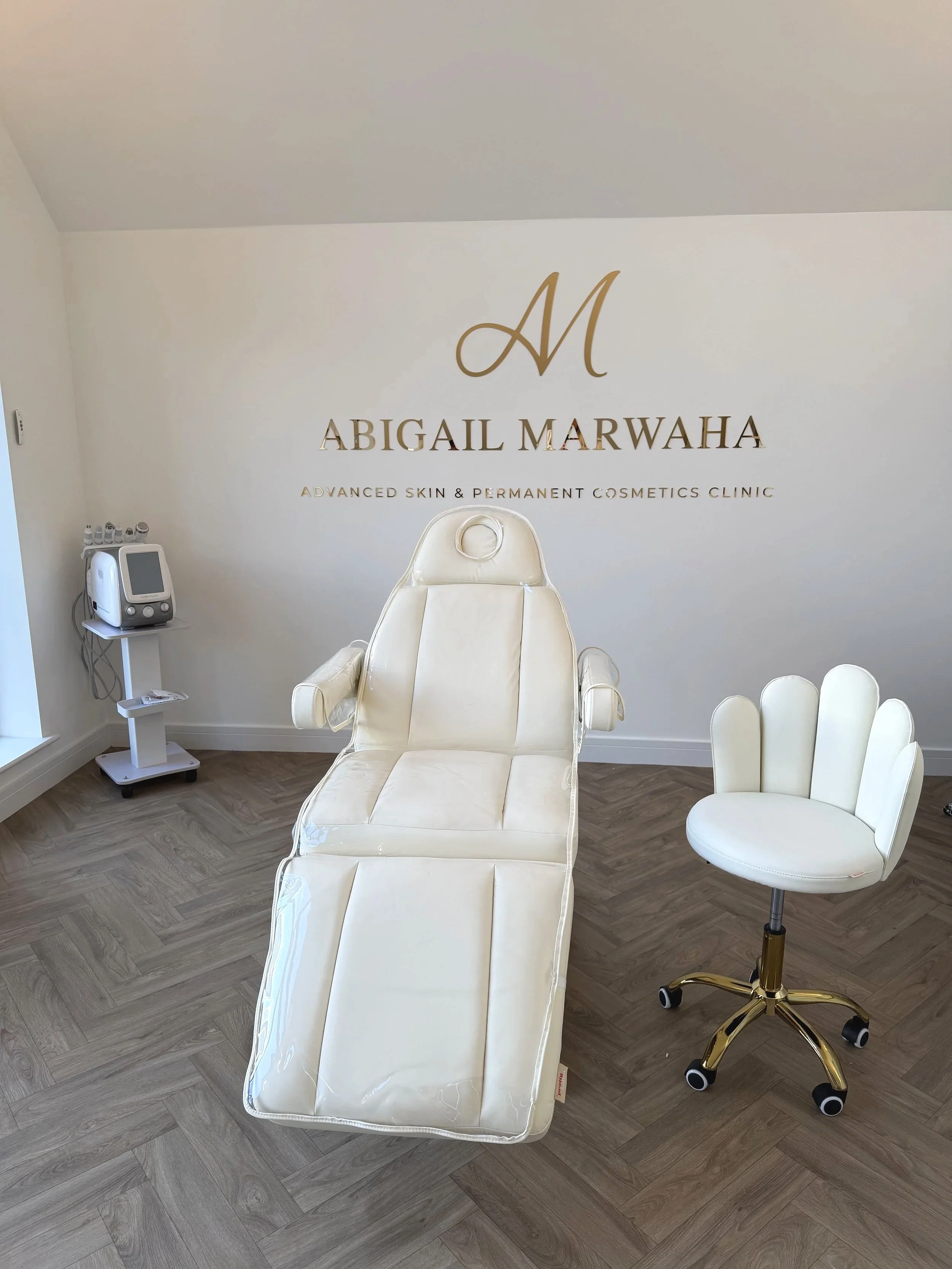 Medical treatment room with a white reclining chair and a white swivel stool, located in front of a wall with the text 'Abigail Marwaha, Advanced Skin & Permanent Cosmetics Clinic' and a large monogram 'A M' logo.
