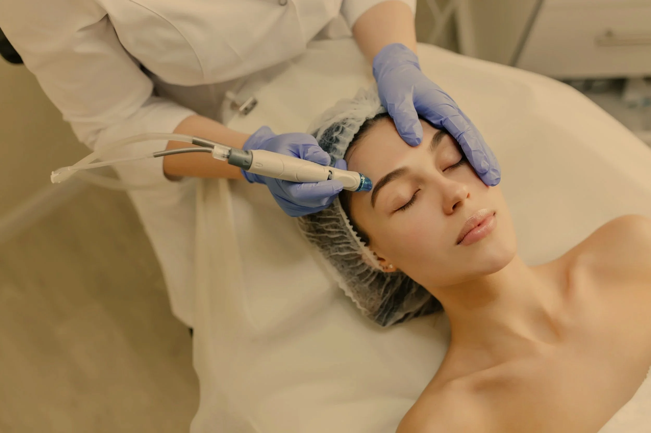 A woman receiving a facial treatment with a handheld device in a spa or clinic setting, lying down with eyes closed, wearing a hair cap and a towel around her shoulders.
