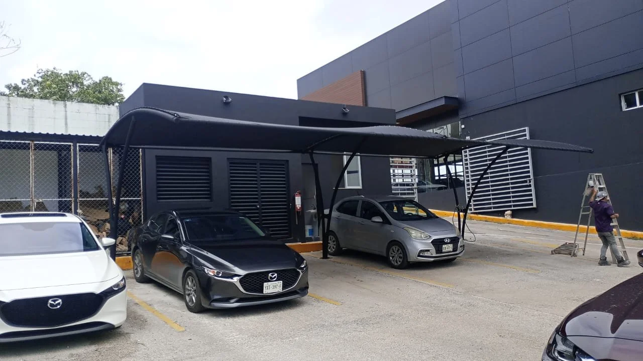 Parking lot with four cars parked under a large black shade structure in front of a modern gray building.