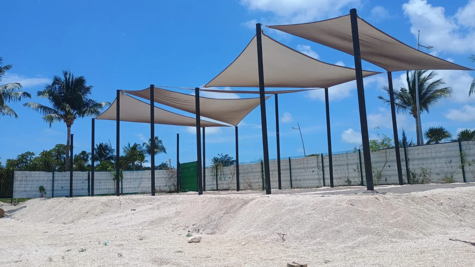Beach with white sand, privacy fence, and large shade sails supported by tall black poles, with a clear blue sky and palm trees in the background.