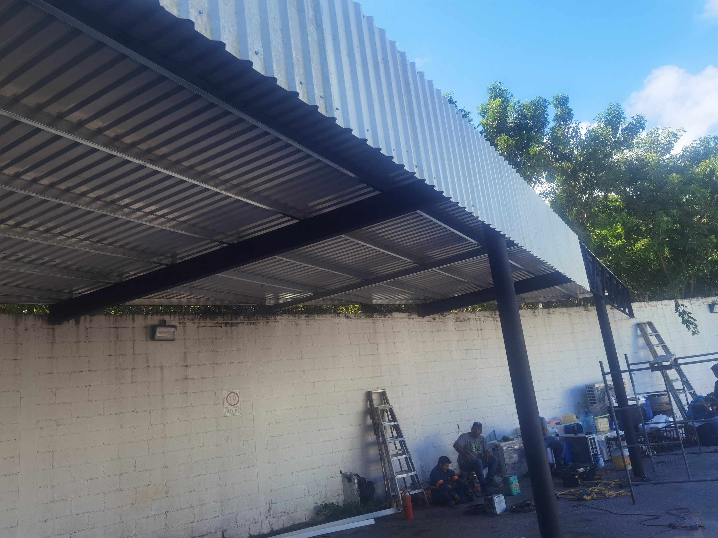 Group of workers sitting and standing under a metal carport structure with ladders, tools, and supplies against a white brick wall, with trees and blue sky in the background.