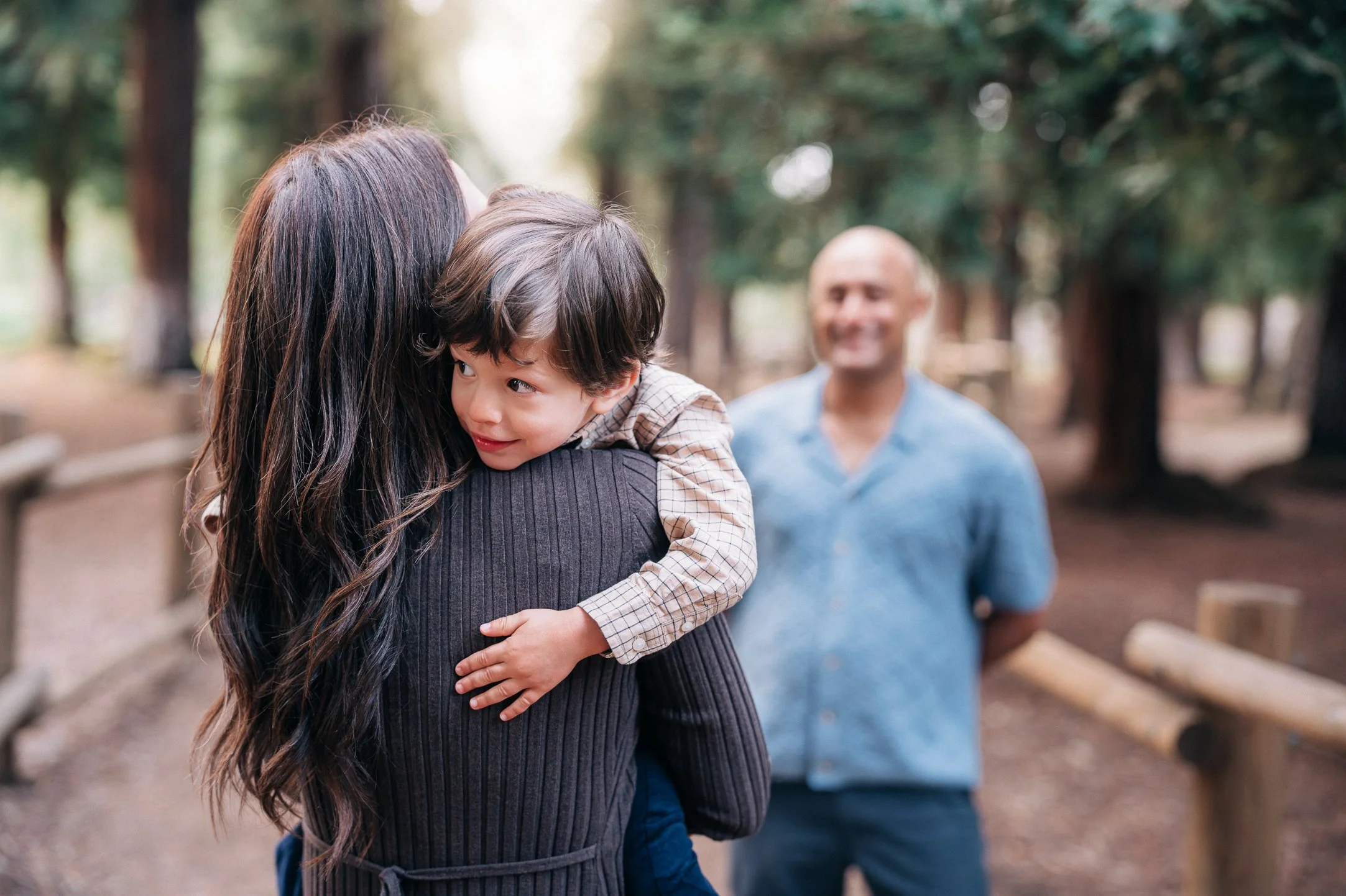 A woman with long dark hair hugging a young boy with dark hair and a plaid shirt, outdoor setting with trees.
