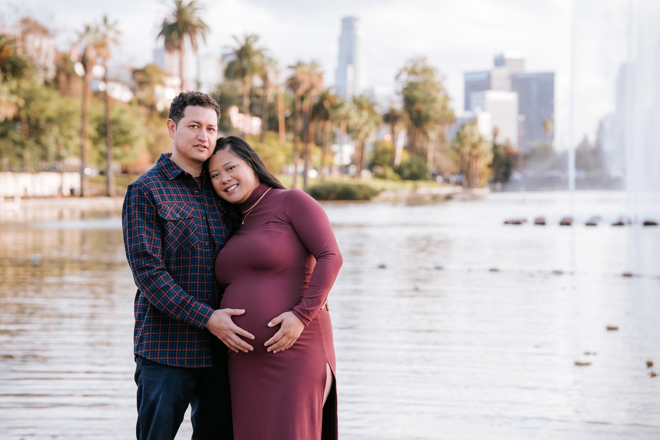 A couple standing by a lake with city buildings and palm trees in the background. The woman is pregnant, and the man has his hand on her belly. They are smiling and looking at the camera.
