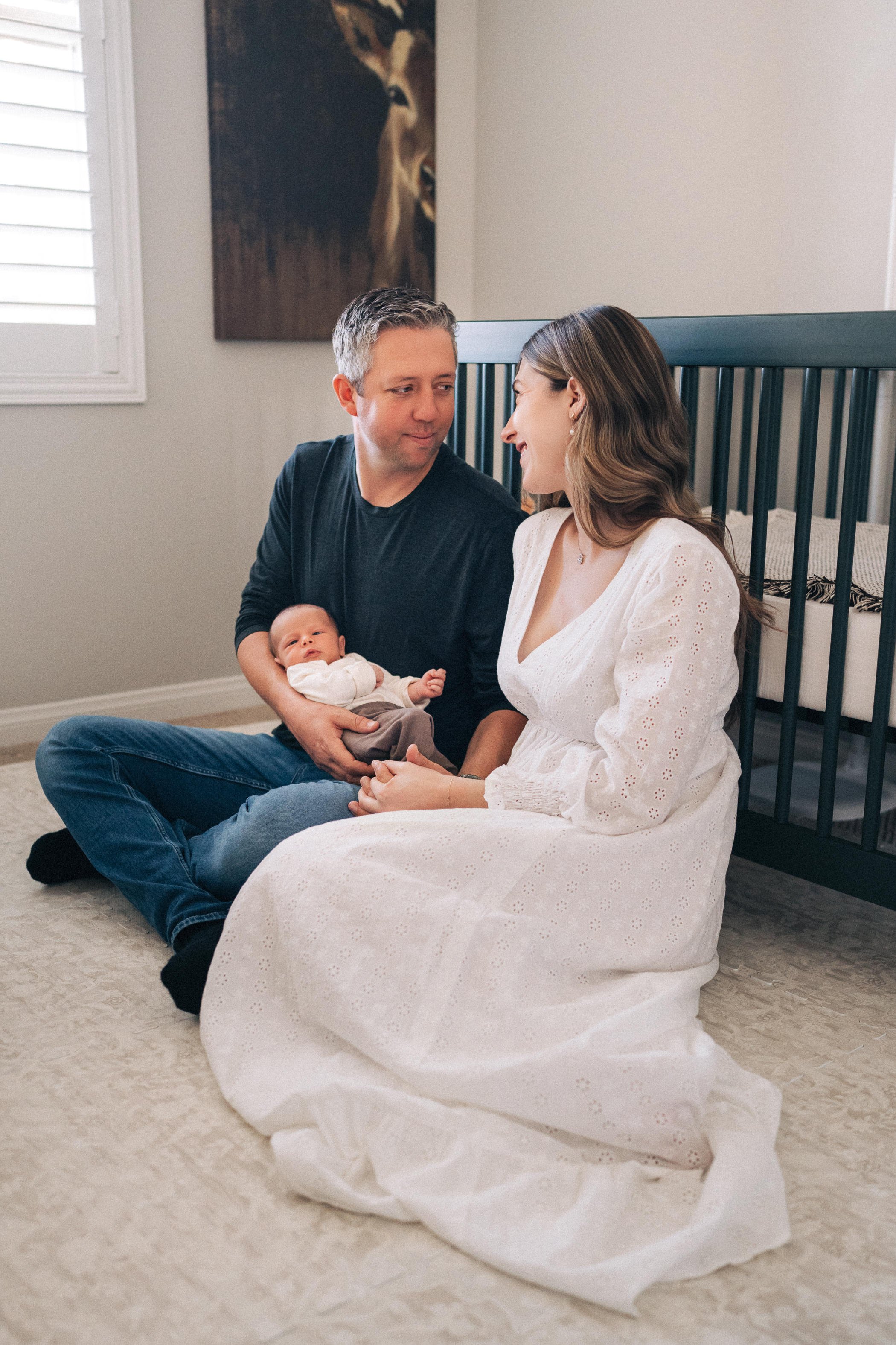 A family sitting on the carpeted floor in a bedroom, with a man holding a baby and a woman sitting close, smiling at each other.