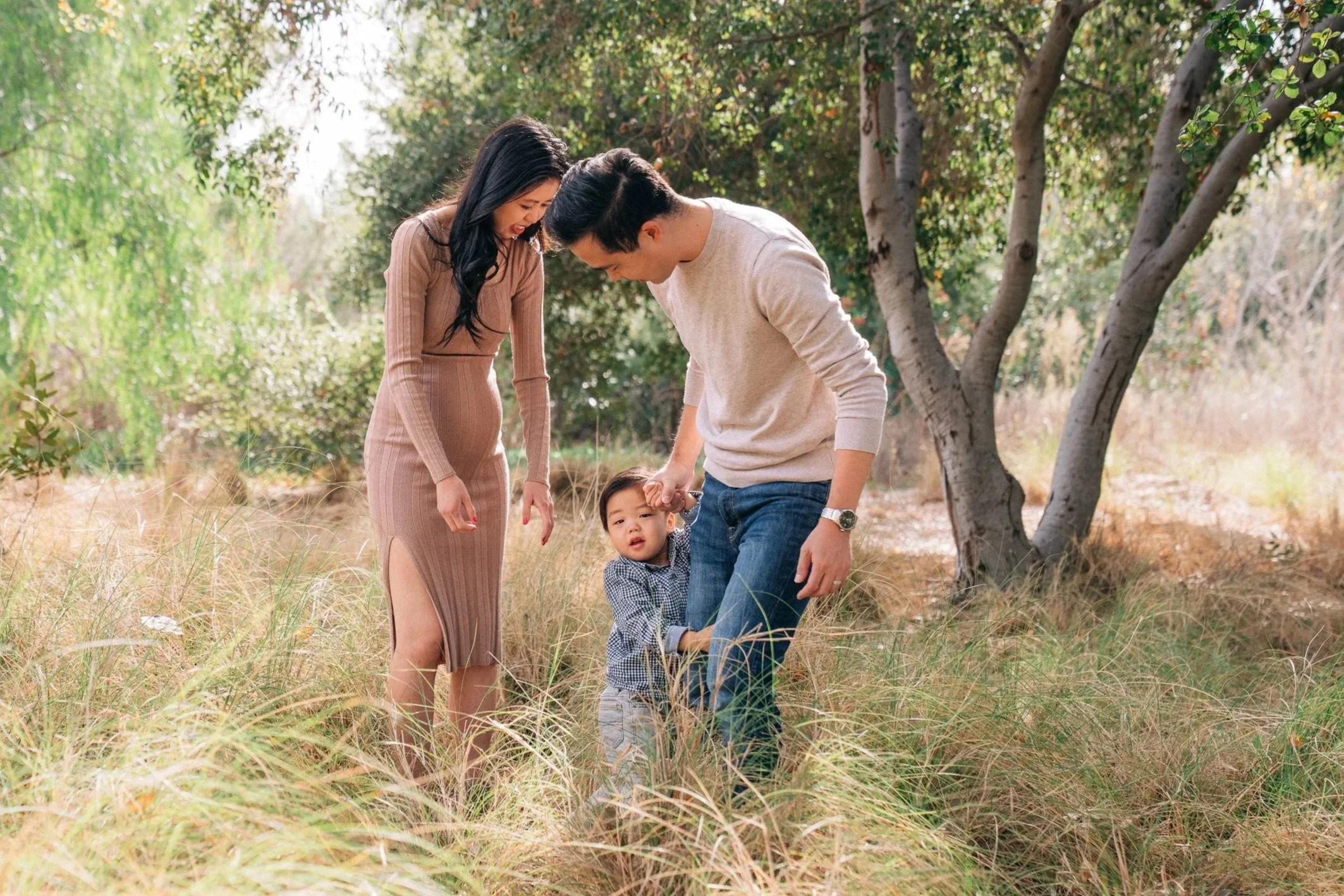 A family of three, a man, a woman, and a young boy, standing outdoors in a grassy area with trees, with the man holding the boy's hand while the boy looks at the camera and the woman watches smiling.