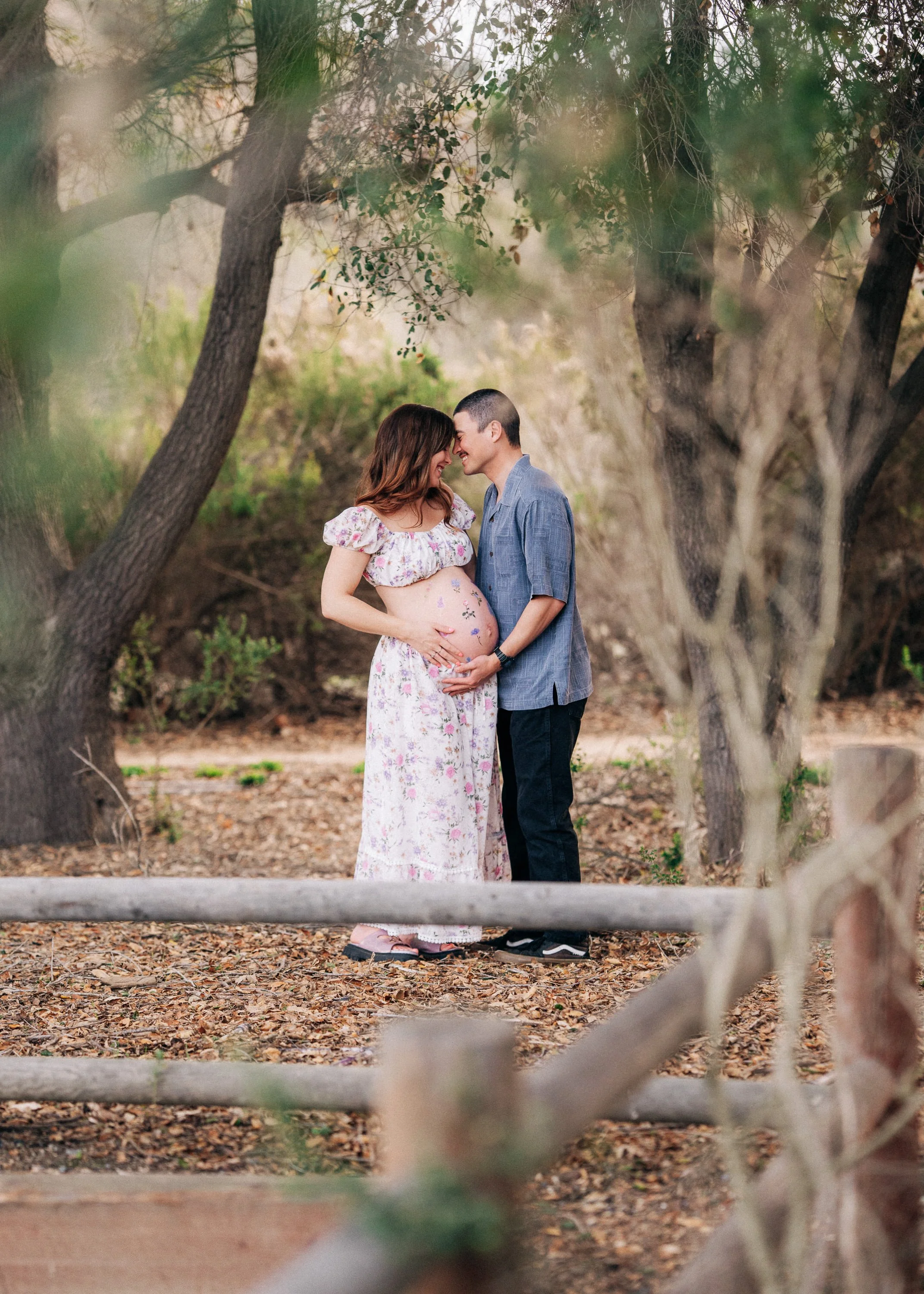 A pregnant woman and a man standing close together in a wooded outdoor setting, touching her belly and smiling at each other.