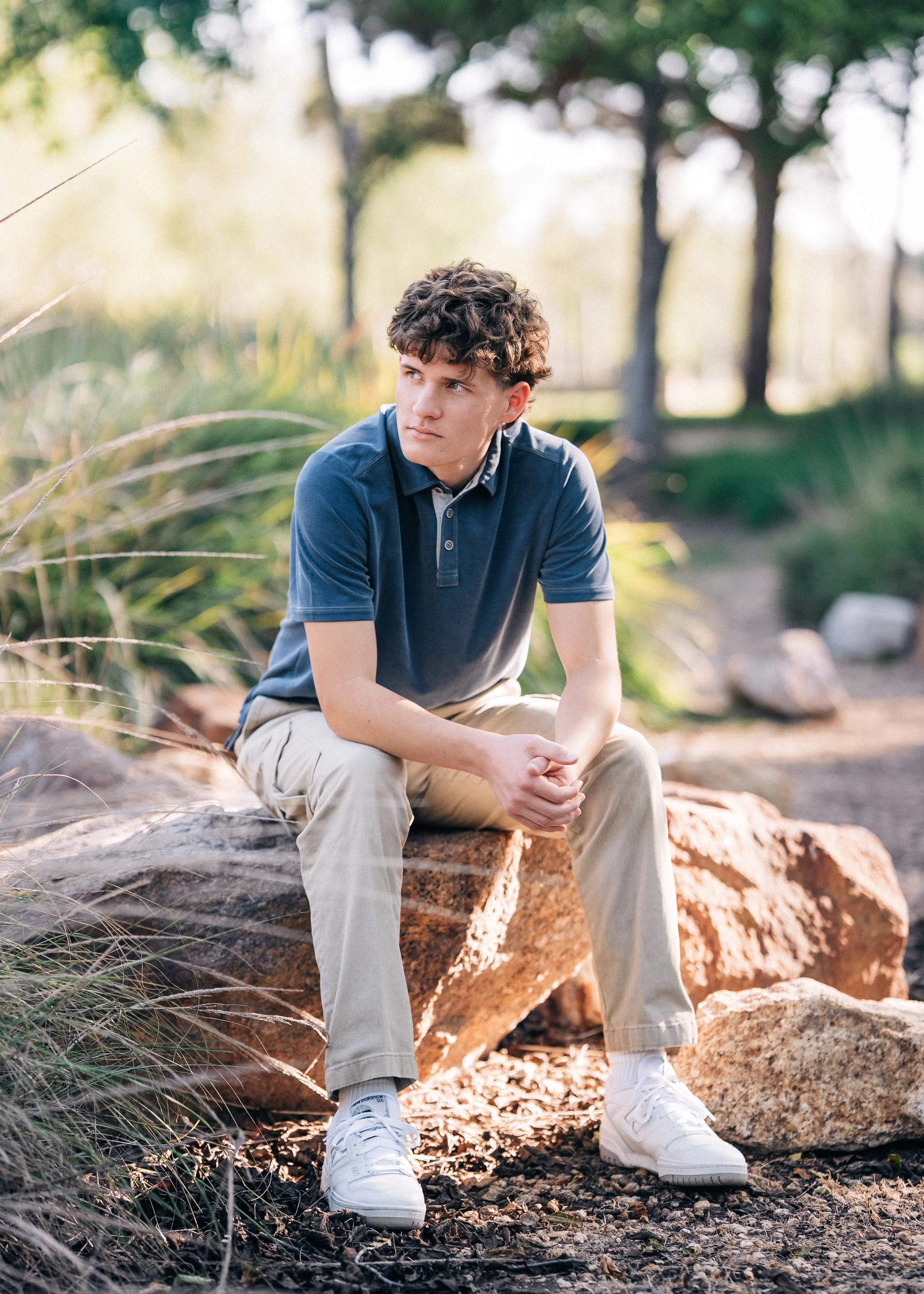 A young man with curly brown hair sitting on a large rock outdoors, wearing a blue polo shirt, khaki pants, and white sneakers, looking thoughtfully to the side with greenery and trees in the background.
