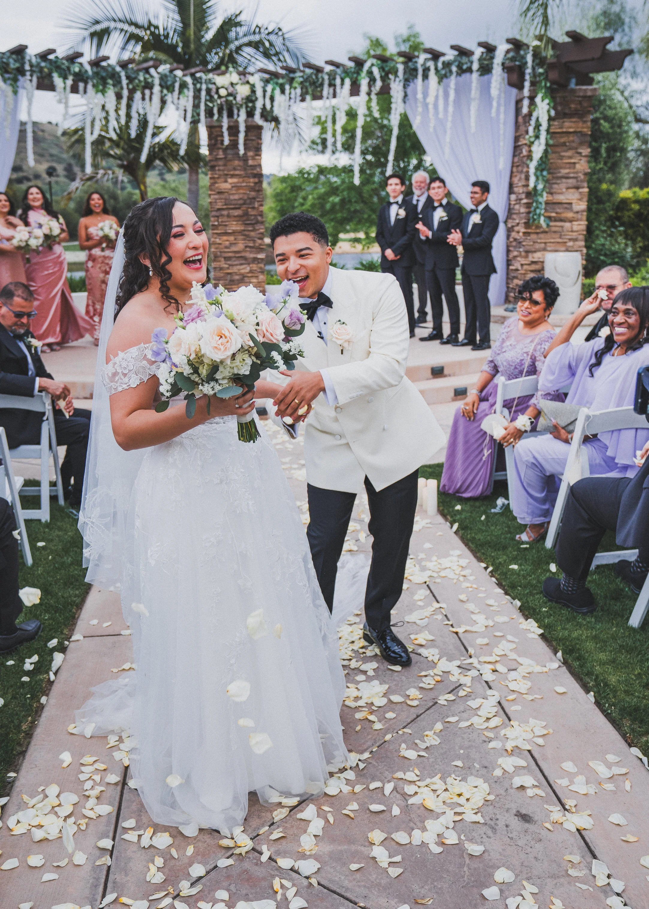 Bride and groom smiling as they walk down the aisle at an outdoor wedding ceremony, surrounded by friends and family, with flower petals on the ground and floral decorations overhead.