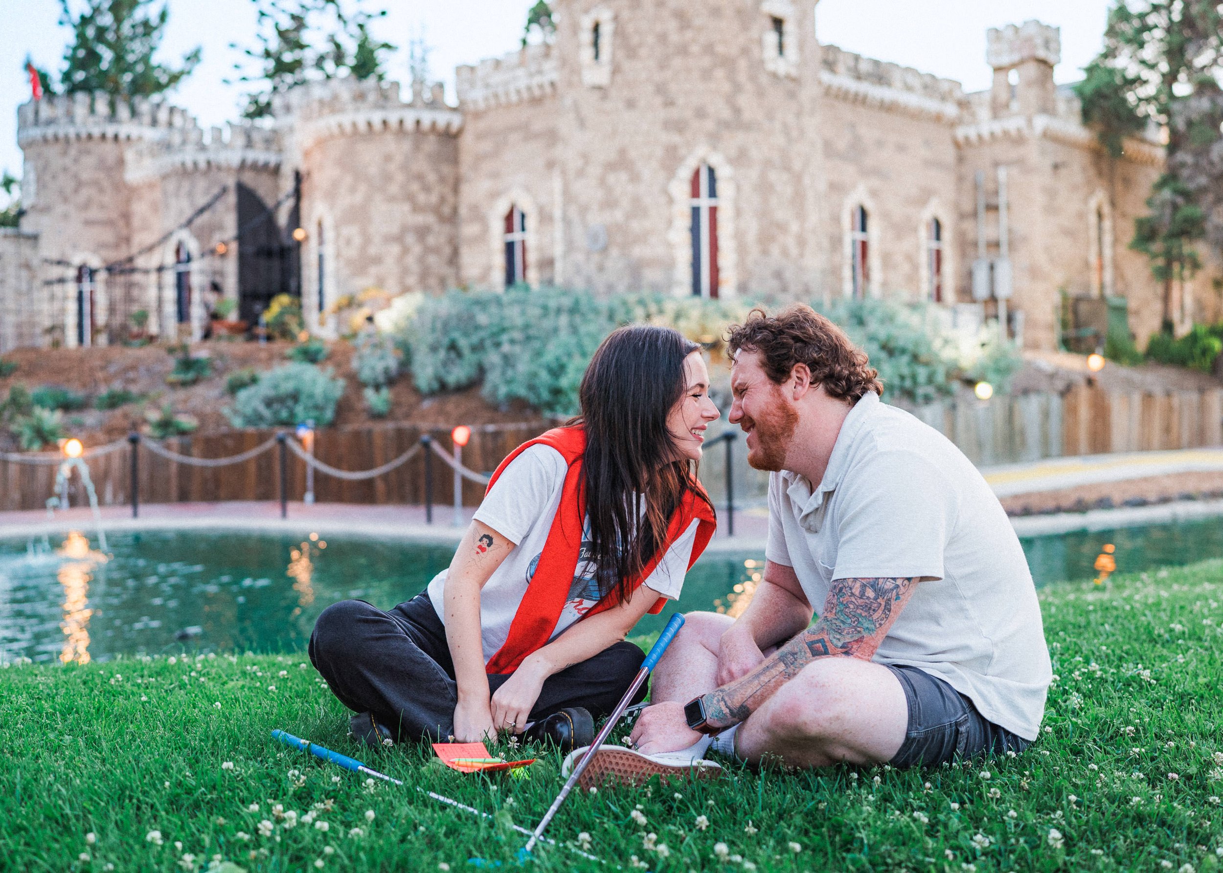 A couple sitting on green grass near a pond, leaning their foreheads together and smiling, with a castle-like building in the background.