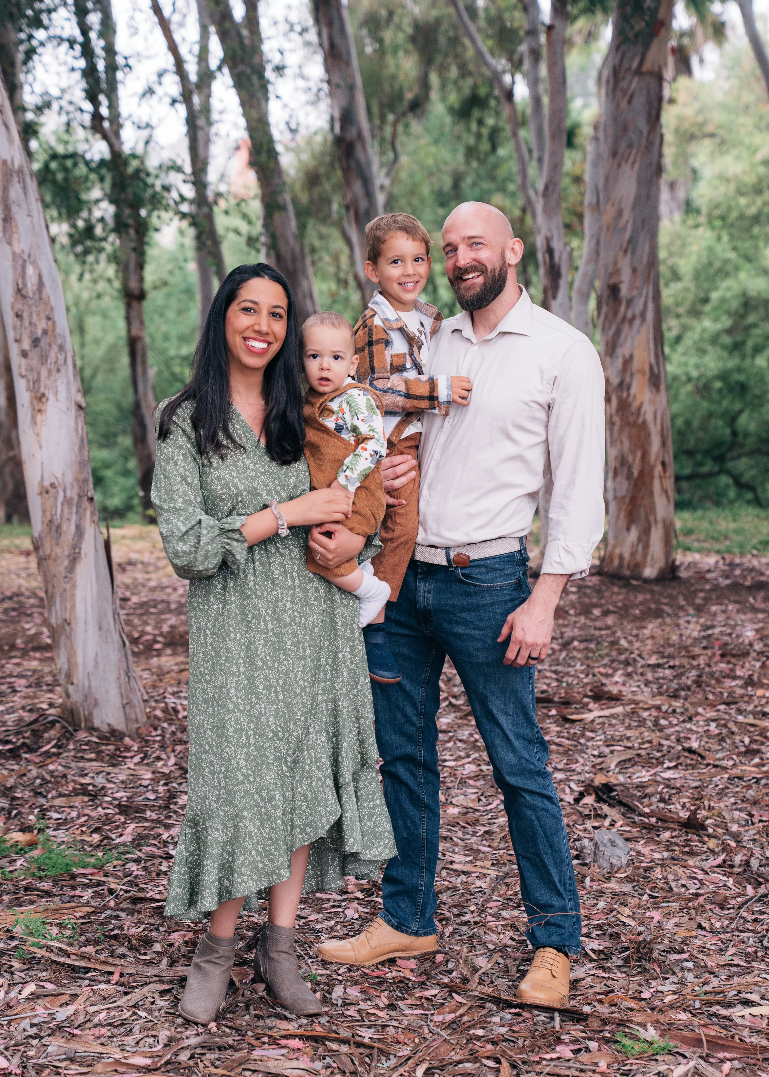 Family photo of four with two children, standing outdoors in a wooded area. The mother has long dark hair, wearing a green dress, and holding a young child. The father has a beard, wearing a white shirt, and holding an older child. All are smiling.