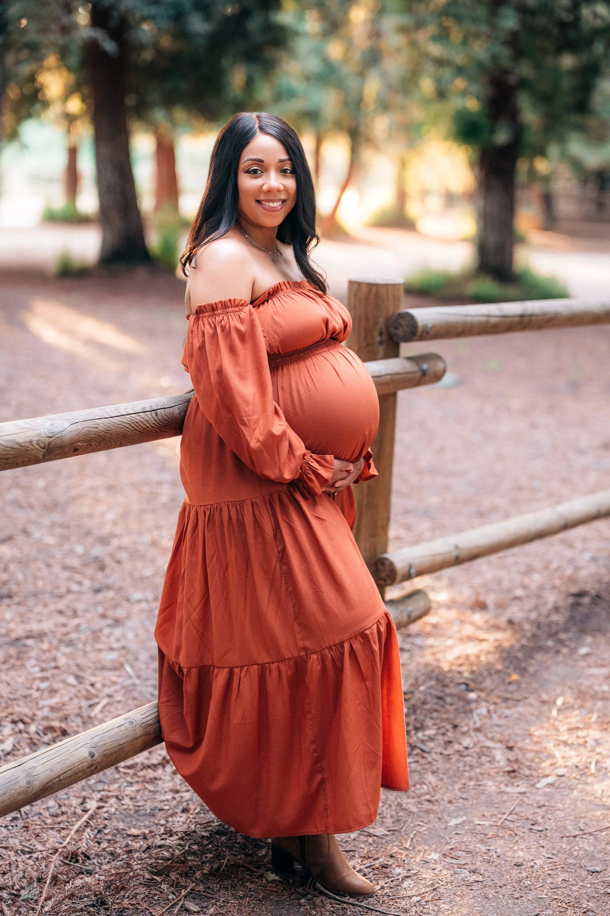 A pregnant woman with dark hair, smiling, standing outdoors by a wooden fence in a park, wearing an off-the-shoulder rust-colored dress and brown boots.