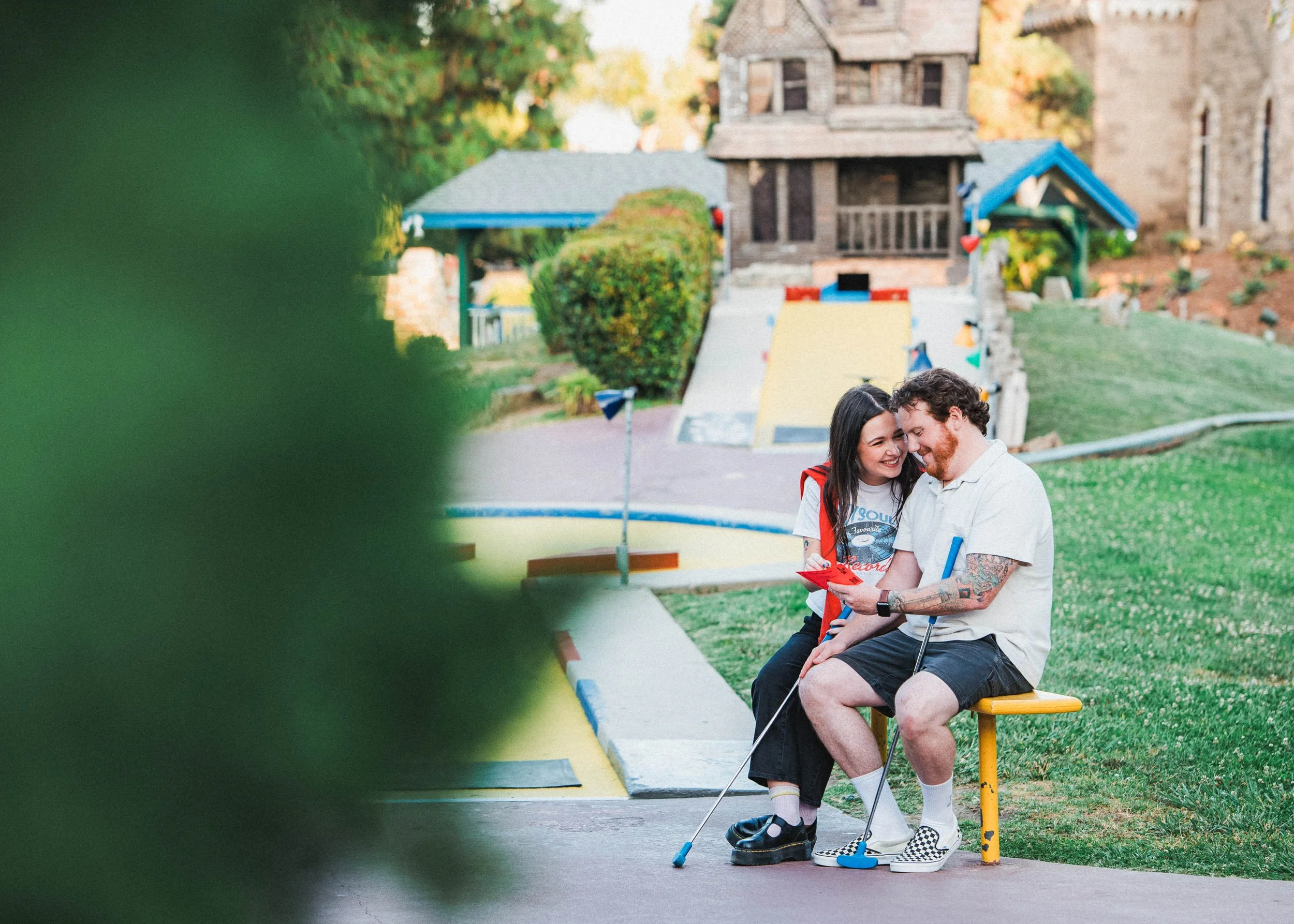 A young woman and a young man sitting together on a yellow bench at a miniature golf course, smiling and looking at a phone, with mini golf structures in the background.