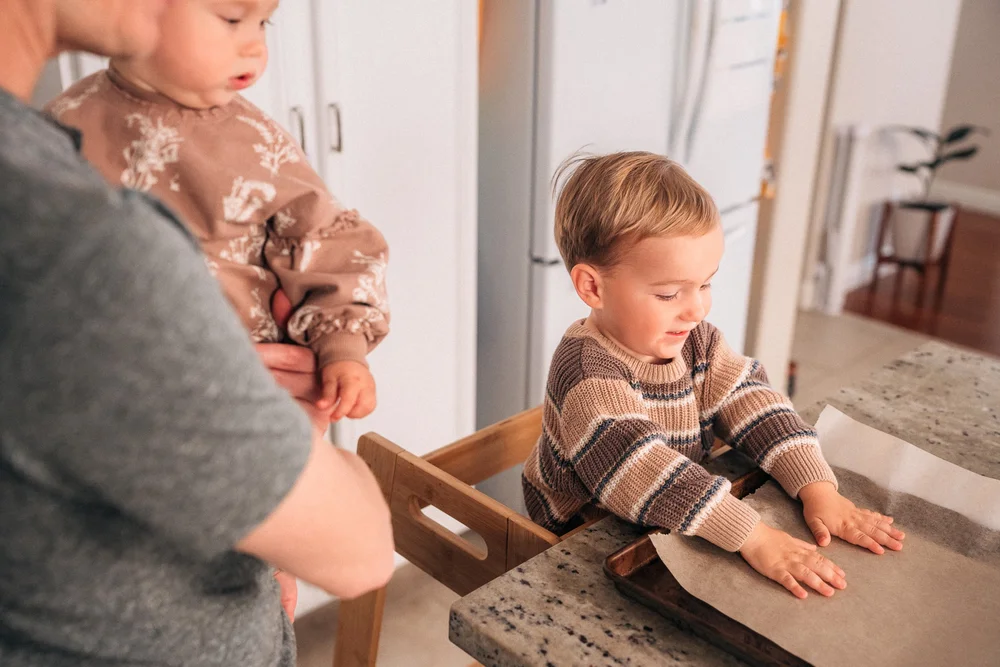 Mom holding baby on her hip while their toddler reaches across the kitchen counter