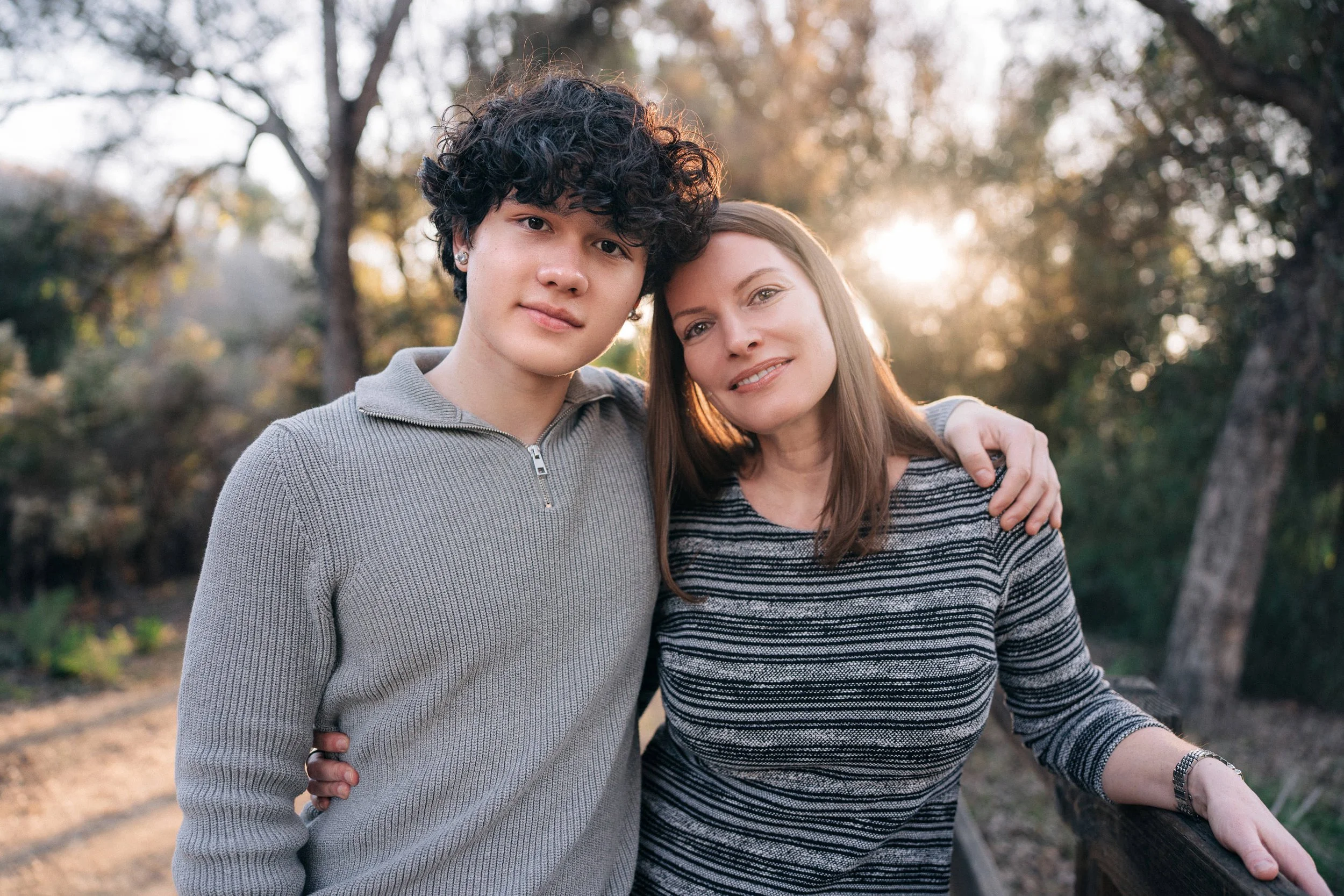 A young man with curly dark hair and a woman with straight brown hair standing close together outdoors, smiling, with trees and sunlight in the background.