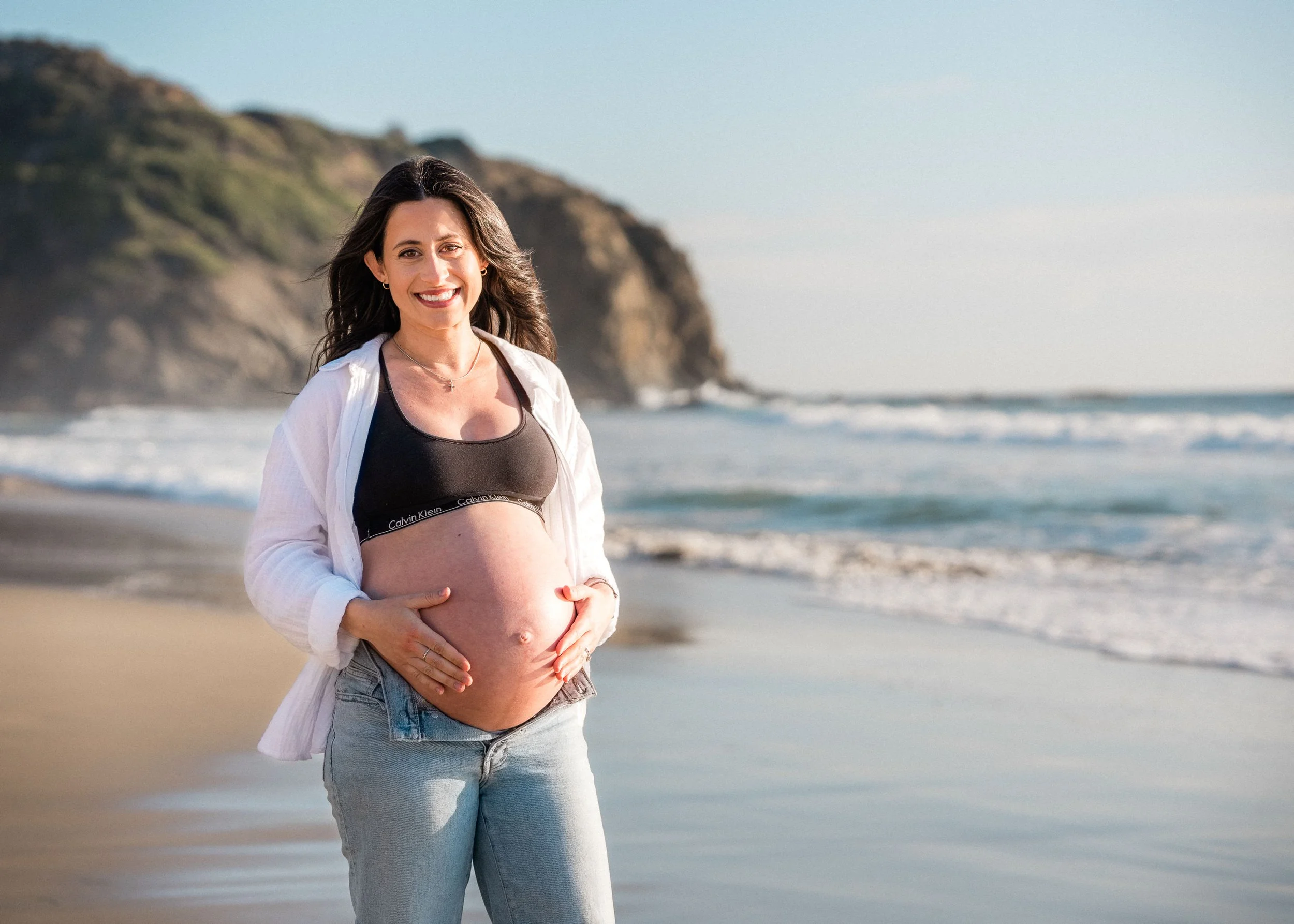 Pregnant woman smiling on the beach, standing with hands on her belly, wearing a black sports bra and light jeans, with cliffs and ocean in the background.