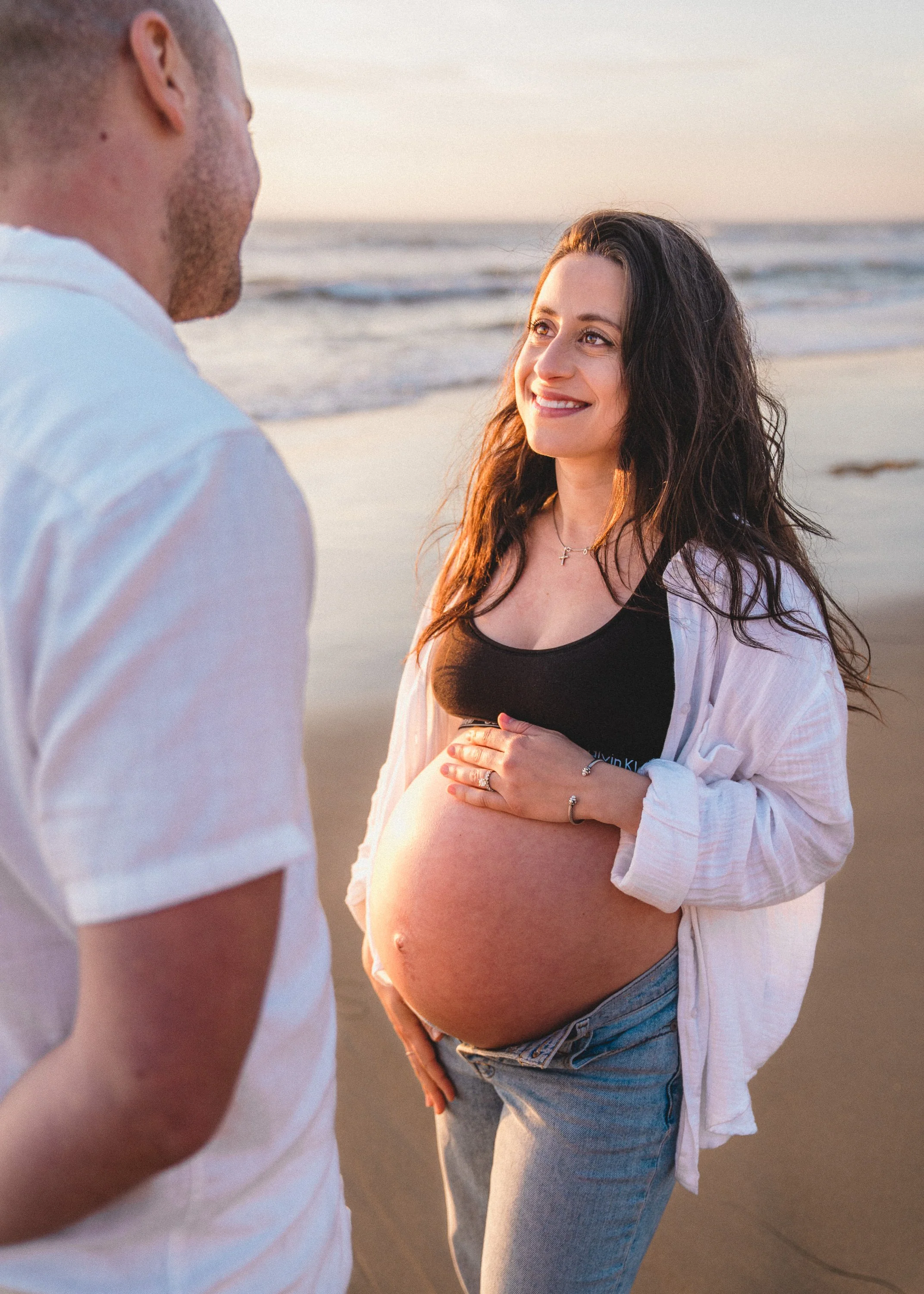 A pregnant woman smiling at her partner on the beach during sunset, holding her belly with one hand while the other rests on her belly, wearing a black top, light jacket, and jeans.