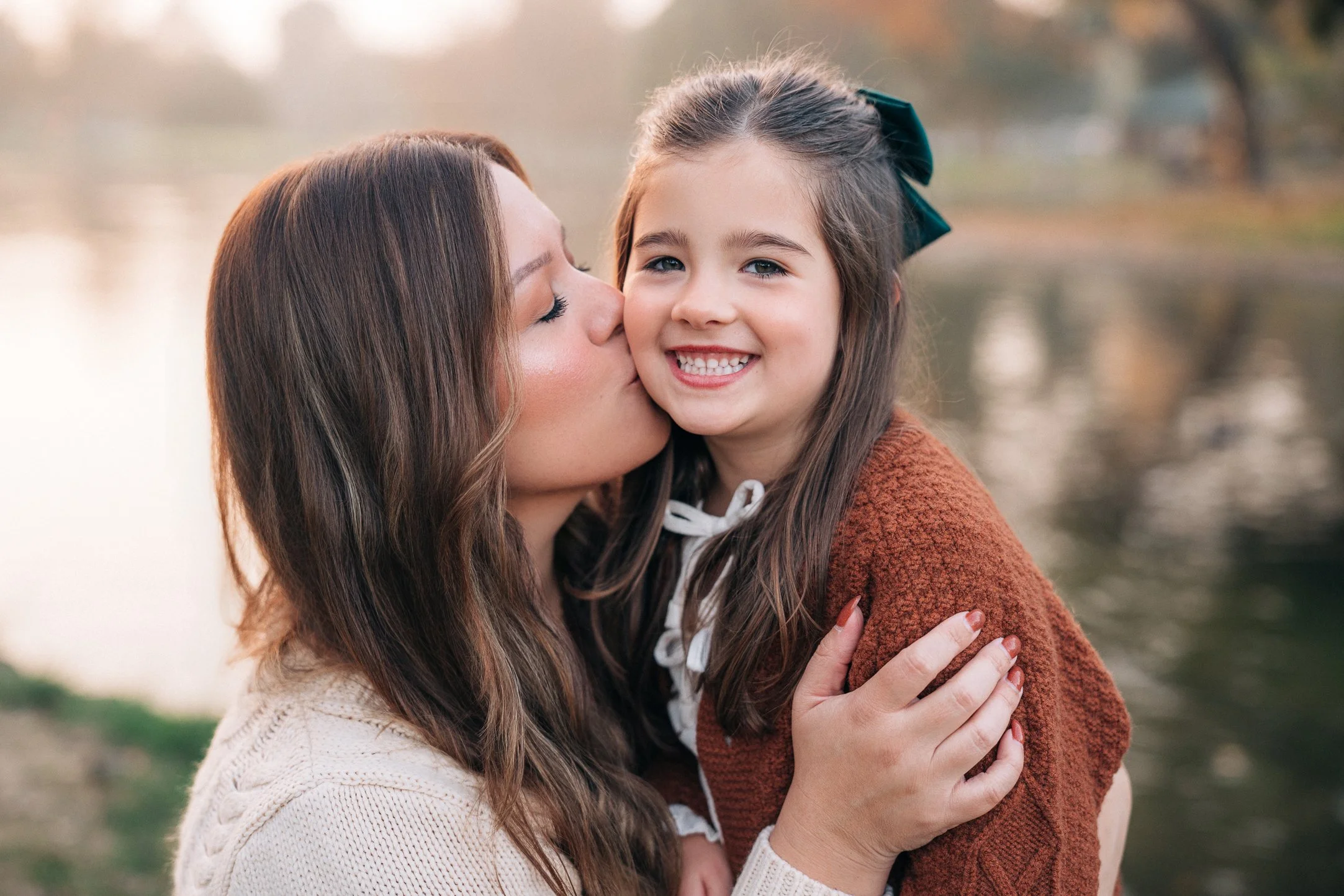 Woman kissing a smiling young girl on the cheek outdoors near a water body during fall.