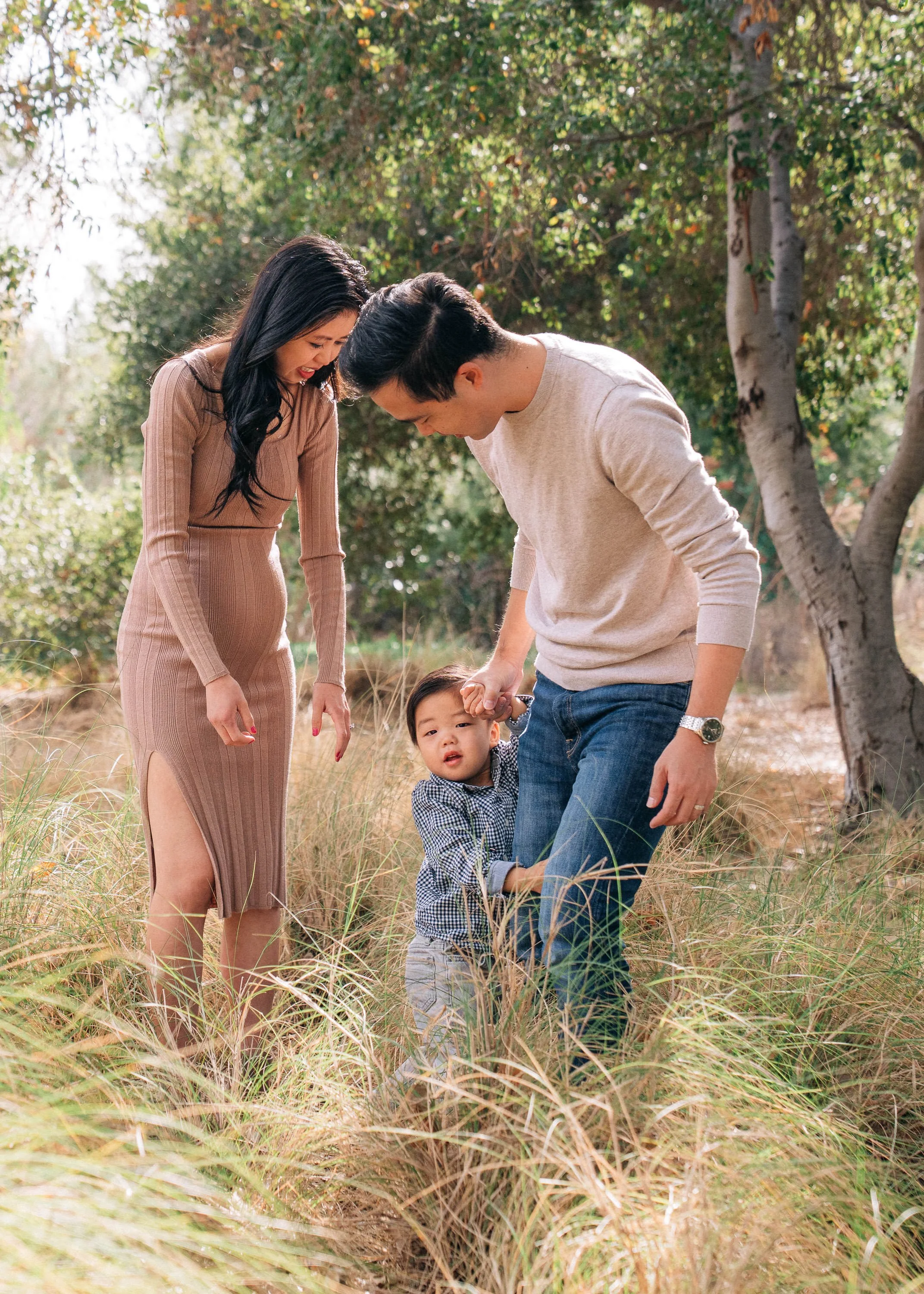 A family of three, including a woman, a man, and a young boy, standing in tall grass in a wooded area. The woman is laughing, the man is looking at the boy, and the boy appears to be upset, clutching the man's leg.