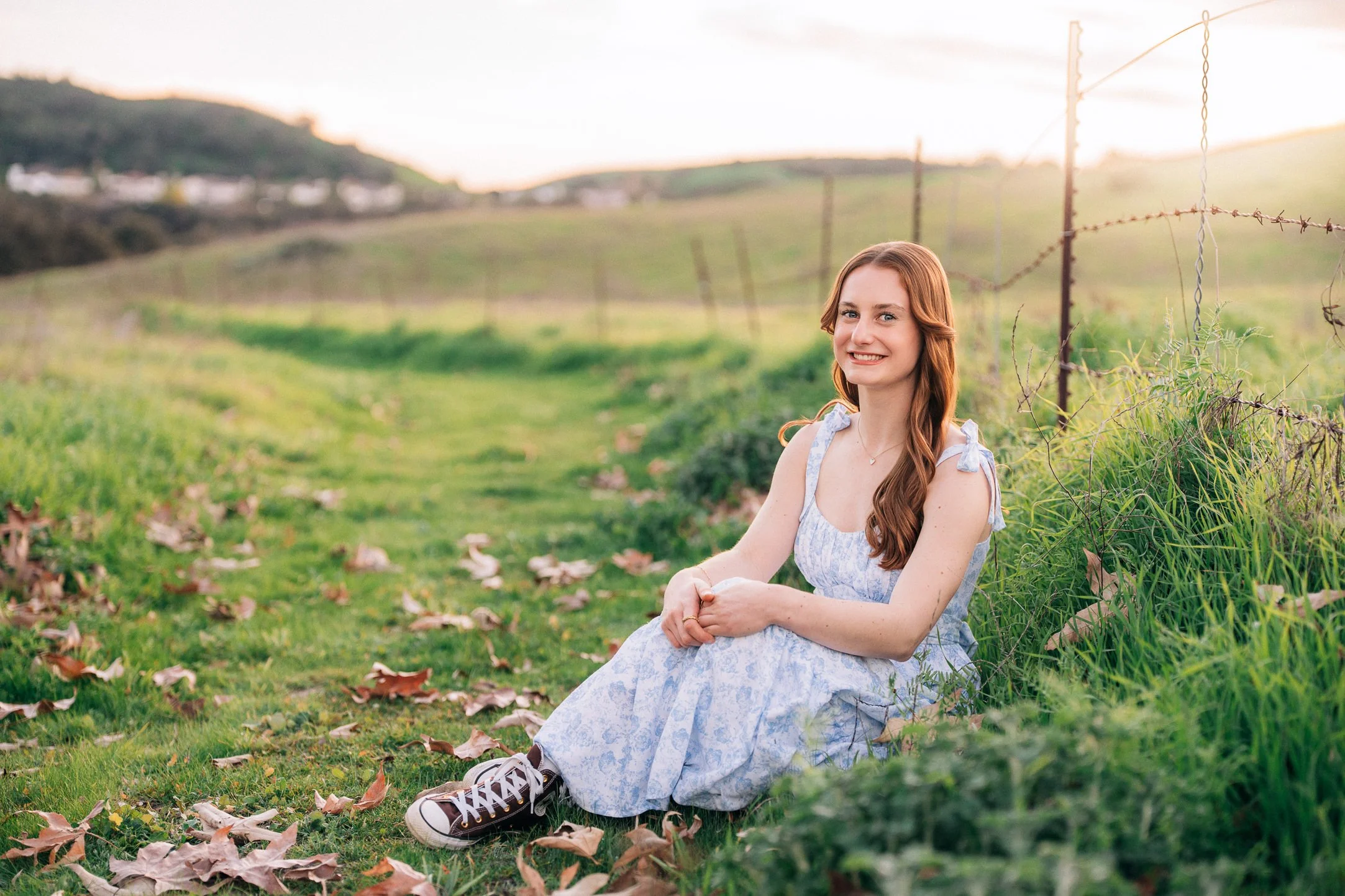 A young woman with long red hair sitting on grass in a field, smiling at the camera during sunset, wearing a light-colored dress and sneakers.