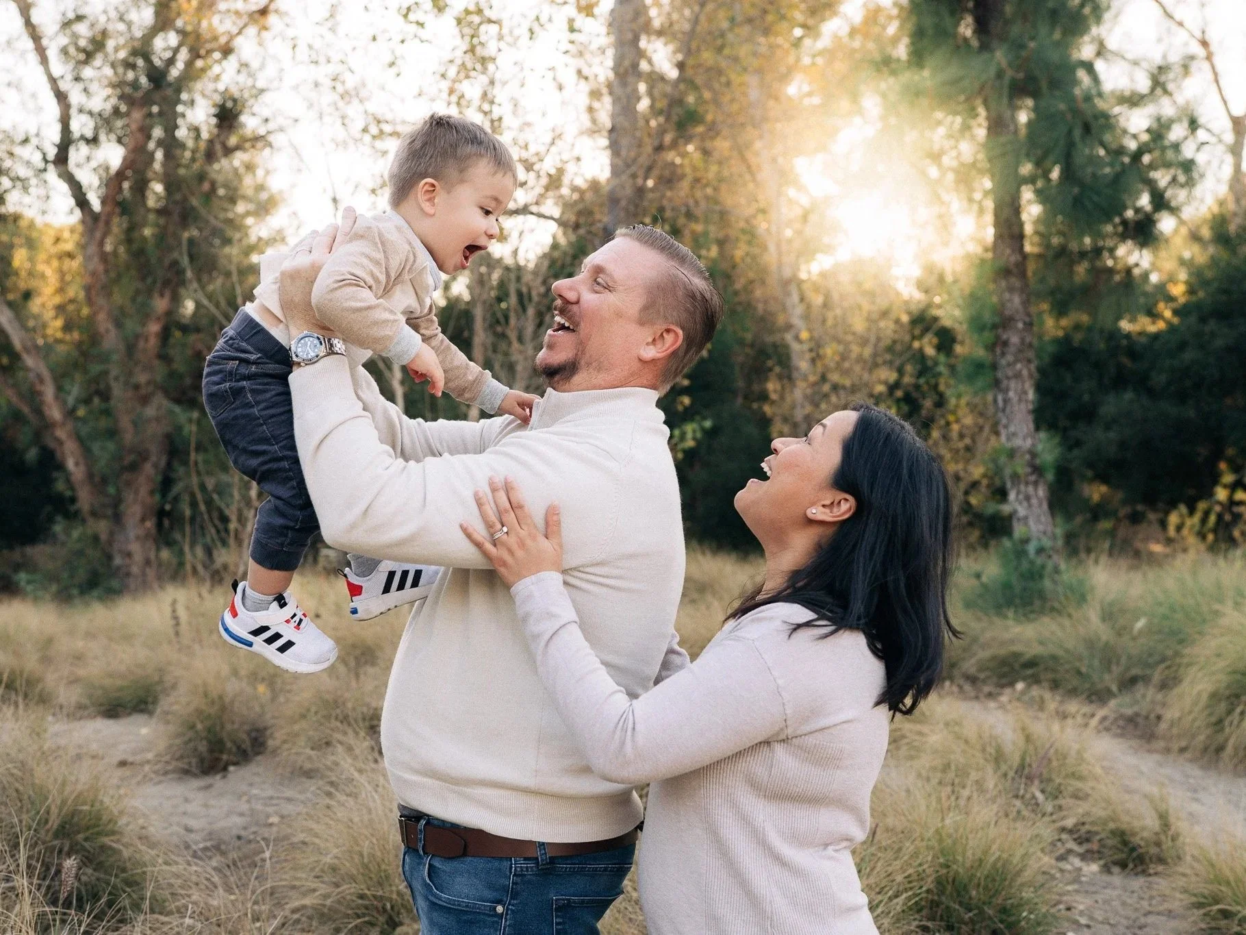 Family of three outdoors on a sunny day; father lifting young boy, mother smiling at them, in a wooded area with trees and grass.