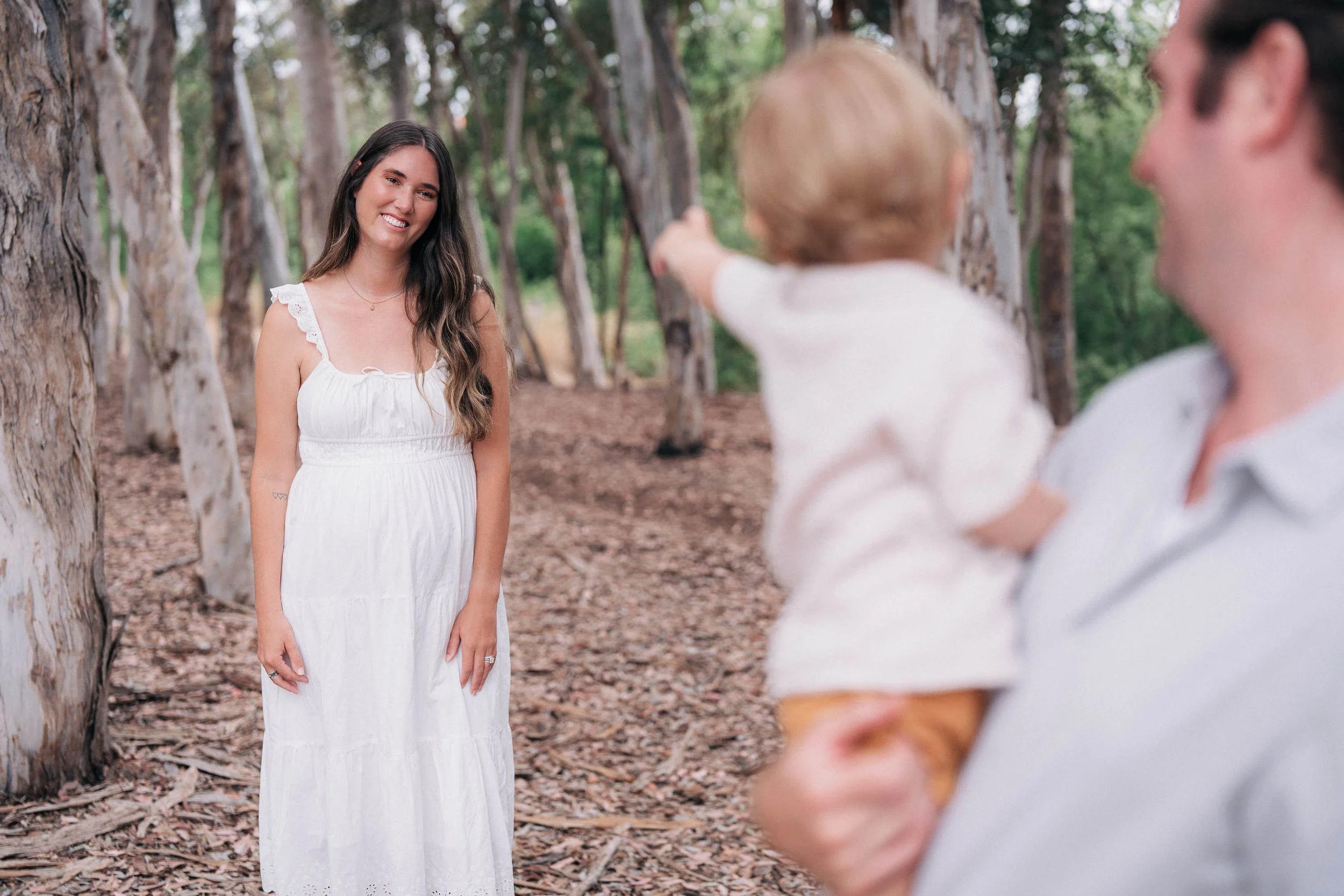 A woman in a white dress standing in a wooded outdoor area, smiling at a man holding a child who is pointing at her.