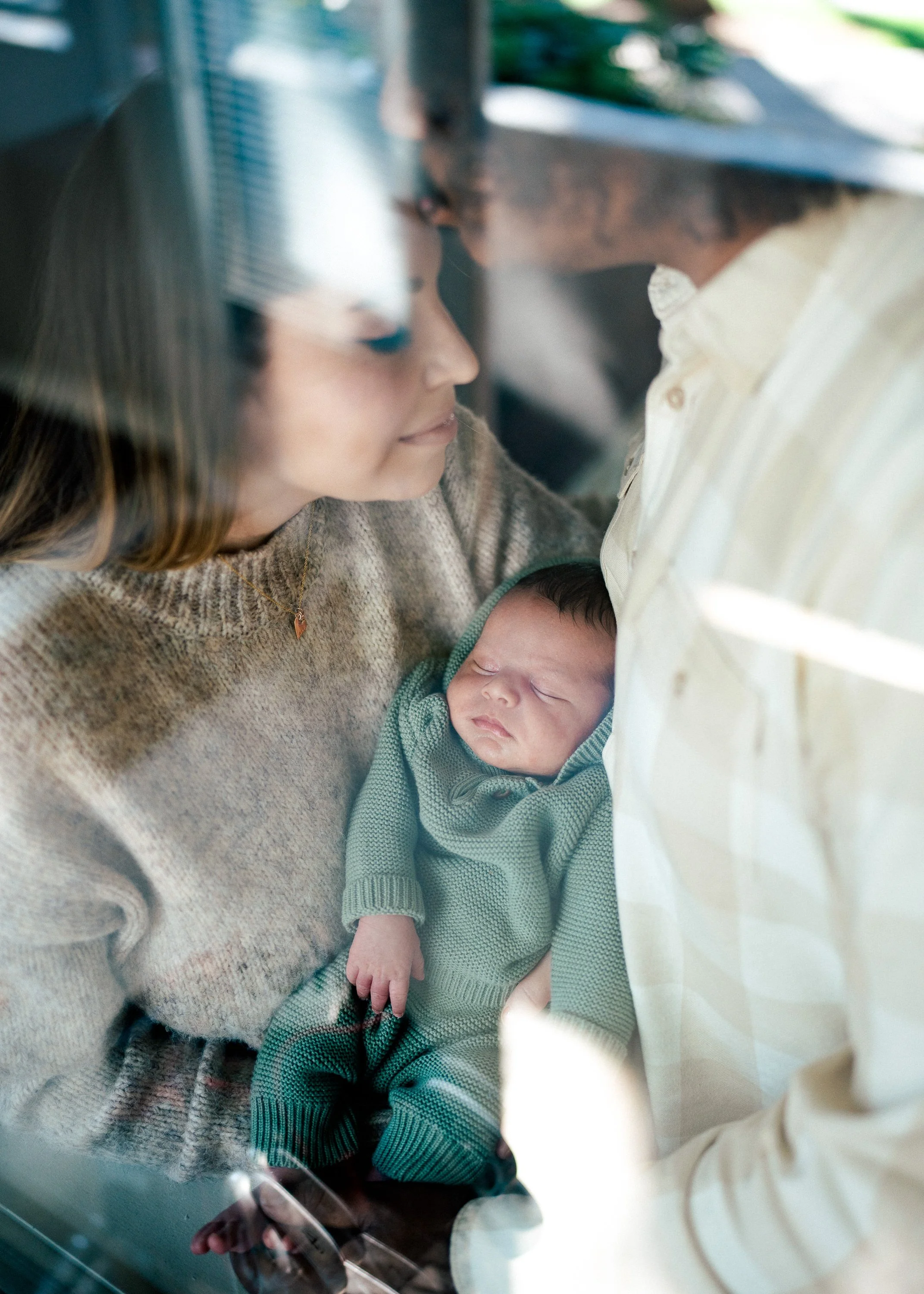 A woman gently holds a sleeping baby wrapped in a green knitted outfit, with both looking at each other through a glass reflection.