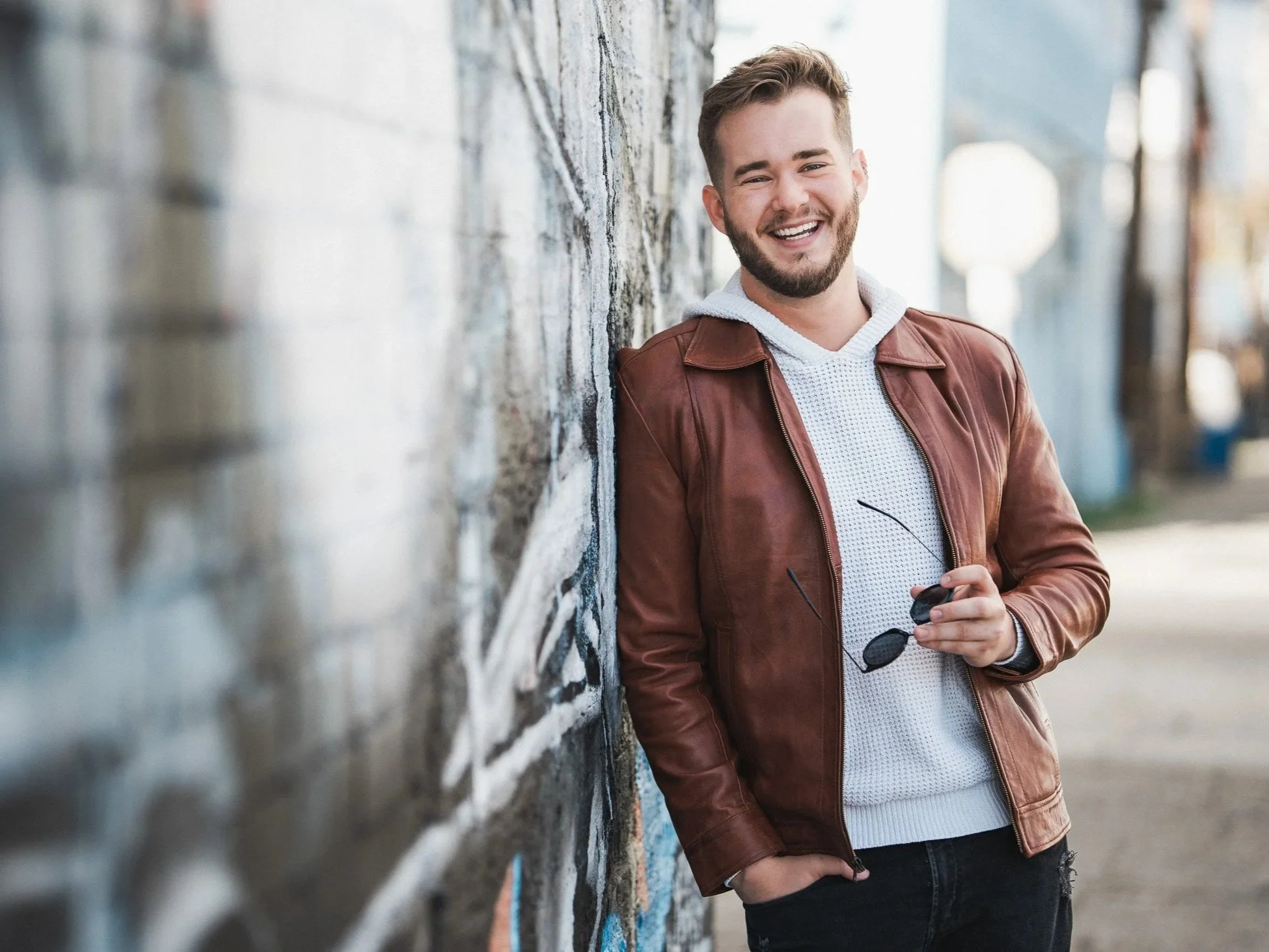 A young man with a beard and short hair smiling and leaning against a brick wall outdoors, wearing a brown leather jacket and holding sunglasses.